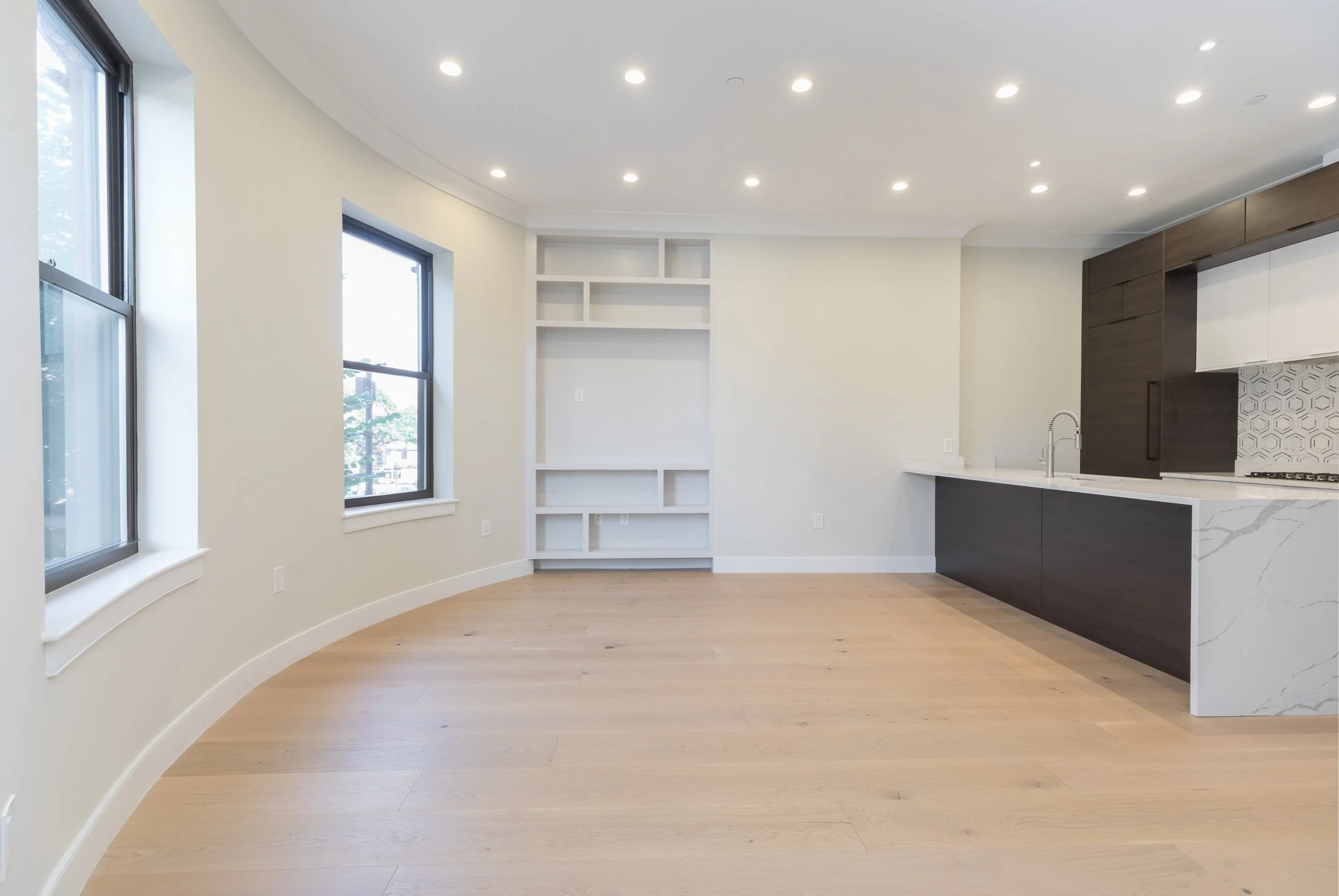 Modern, minimally decorated kitchen with white walls, a built-in white shelving unit, large windows, and a black and white kitchen island with a marble countertop.