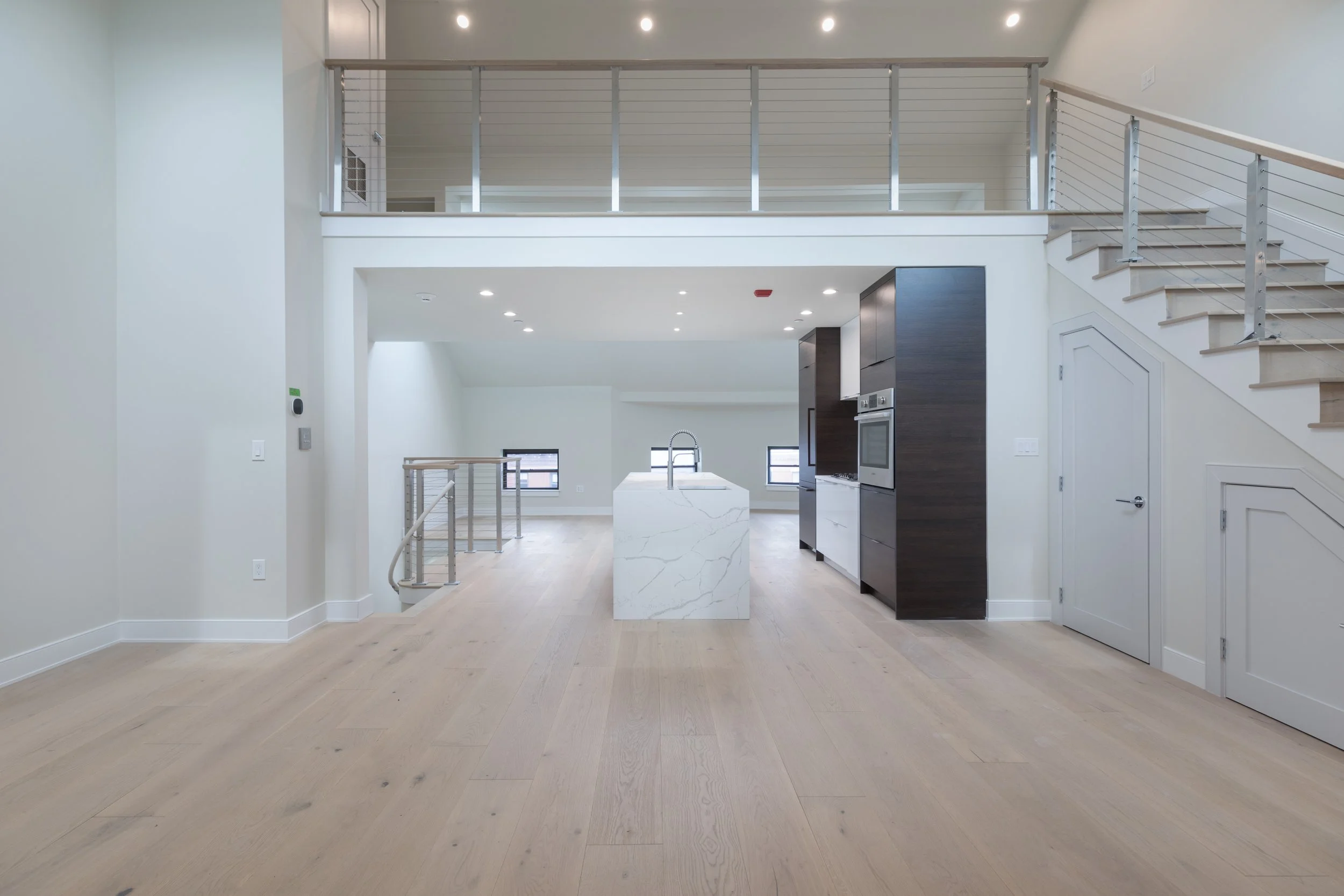 Interior view of a modern, open-concept living area with light wooden flooring, white walls, and a staircase with metal railing leading to an upper level. The space includes a kitchen with a white marble island, dark wood cabinetry, and built-in appl