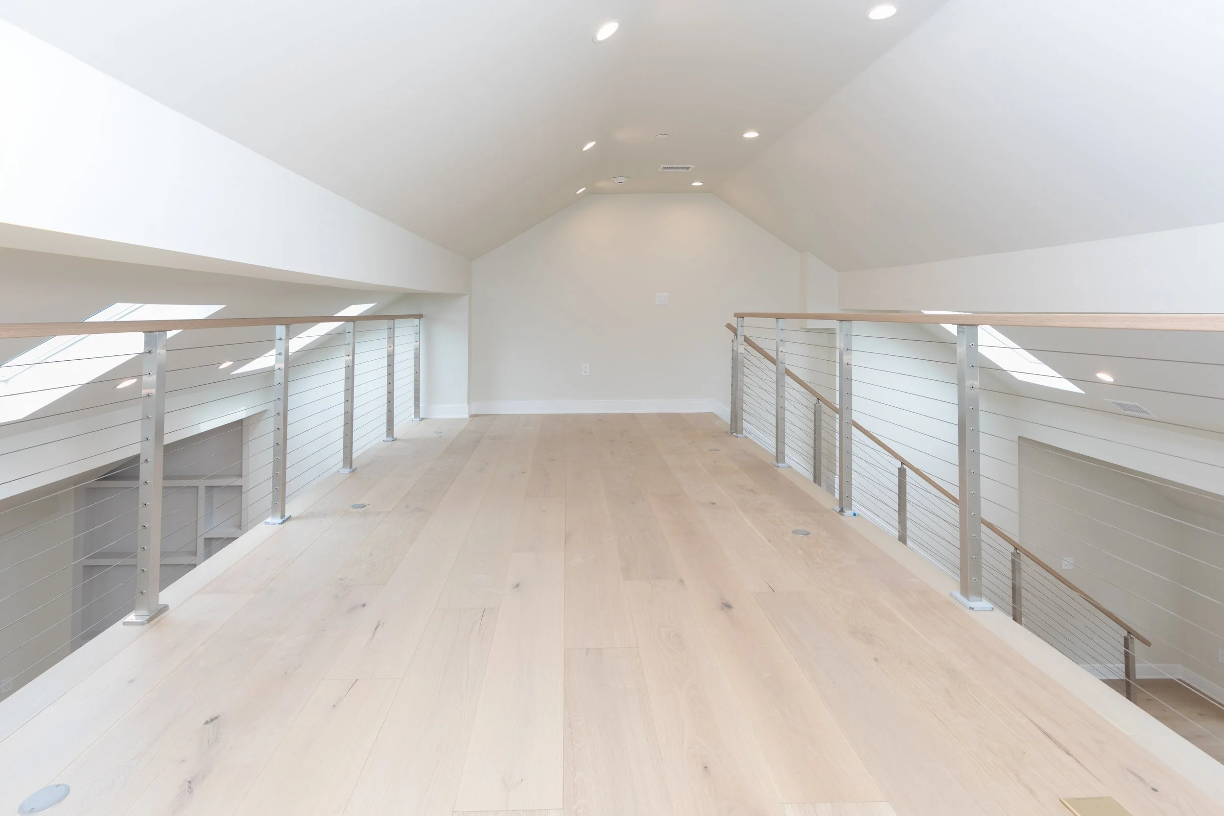 Empty, modern loft with white walls and ceilings, wood flooring, and metal railing, with natural light coming through skylights.