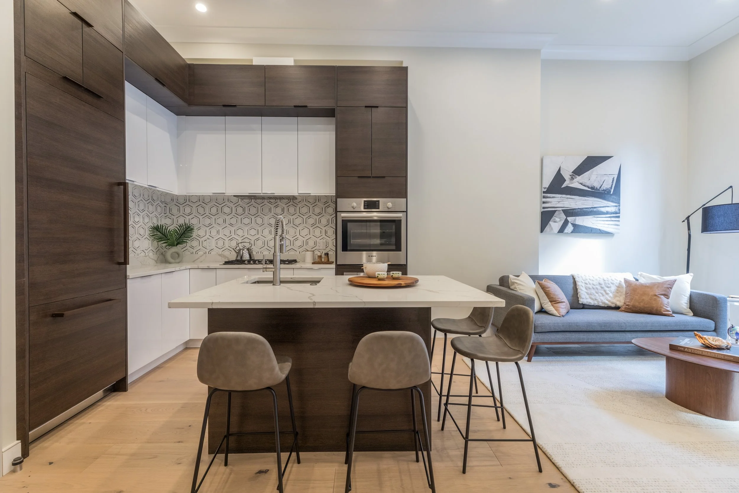 Modern kitchen with white and dark wood cabinets, geometric backsplash, kitchen island with a bowl and utensils, open living area with gray sofa, artwork, and floor lamp.