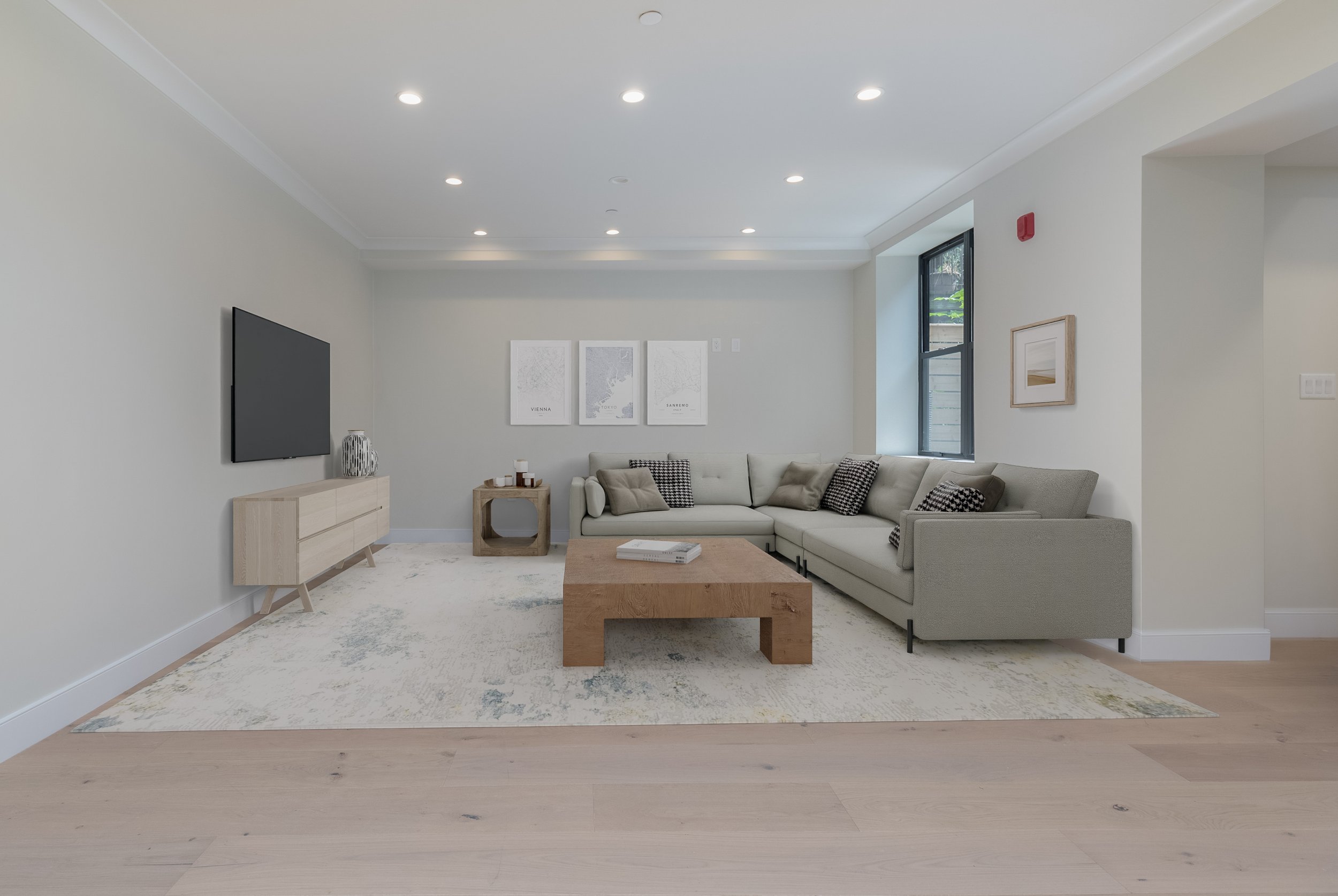 A modern living room with a light-colored sectional sofa, a wooden coffee table, a wall-mounted flat-screen TV, and minimalist artwork on the wall. Large windows let in natural light, and a neutral rug covers part of the wooden floor.