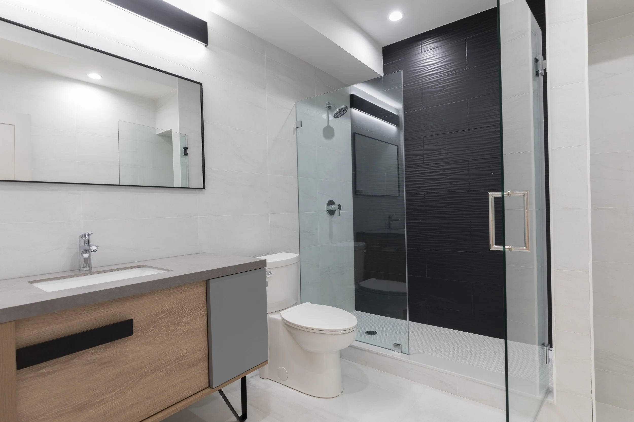 Modern bathroom with white tiled walls, a sleek vanity with a sink, and a glass-enclosed shower area with black textured tiles.