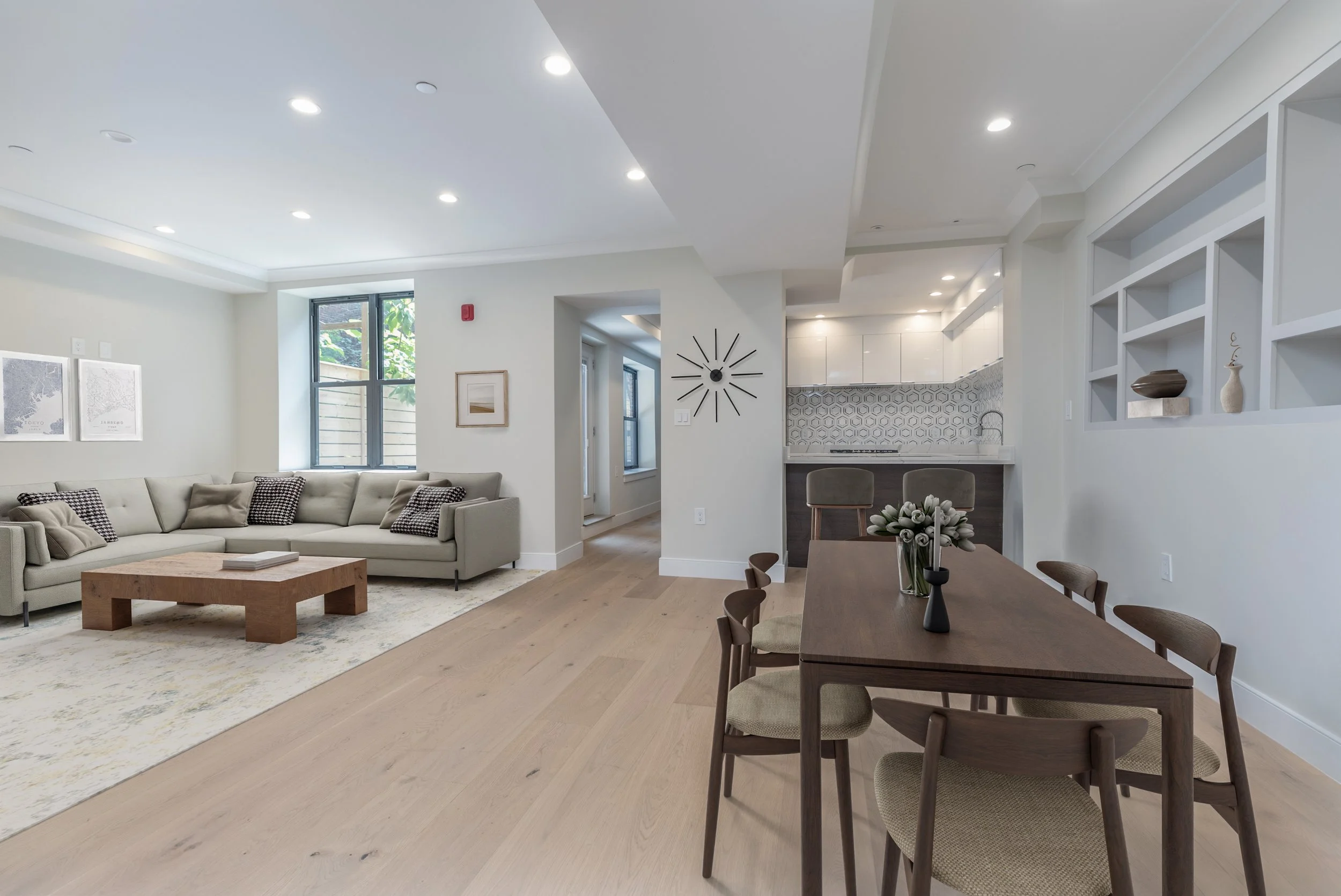 A modern open-concept living room with a beige L-shaped sofa, a wooden coffee table, and a large window with greenery outside. The space includes a dining table with chairs and a kitchen with white cabinets and geometric backsplash. The ceiling has r