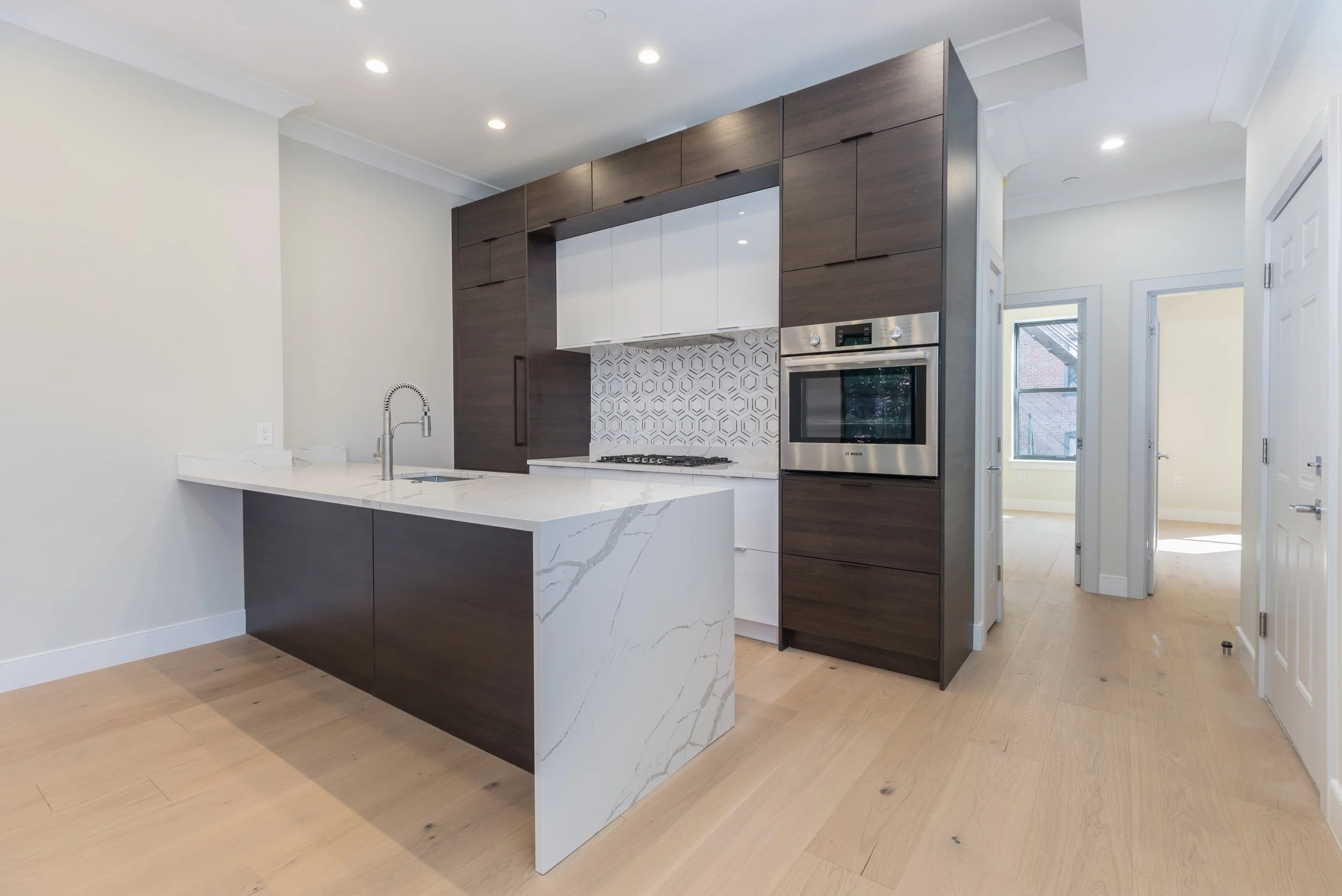 Modern kitchen with white marble island, dark wood cabinets, white backsplash, stainless steel oven, and light hardwood floors.