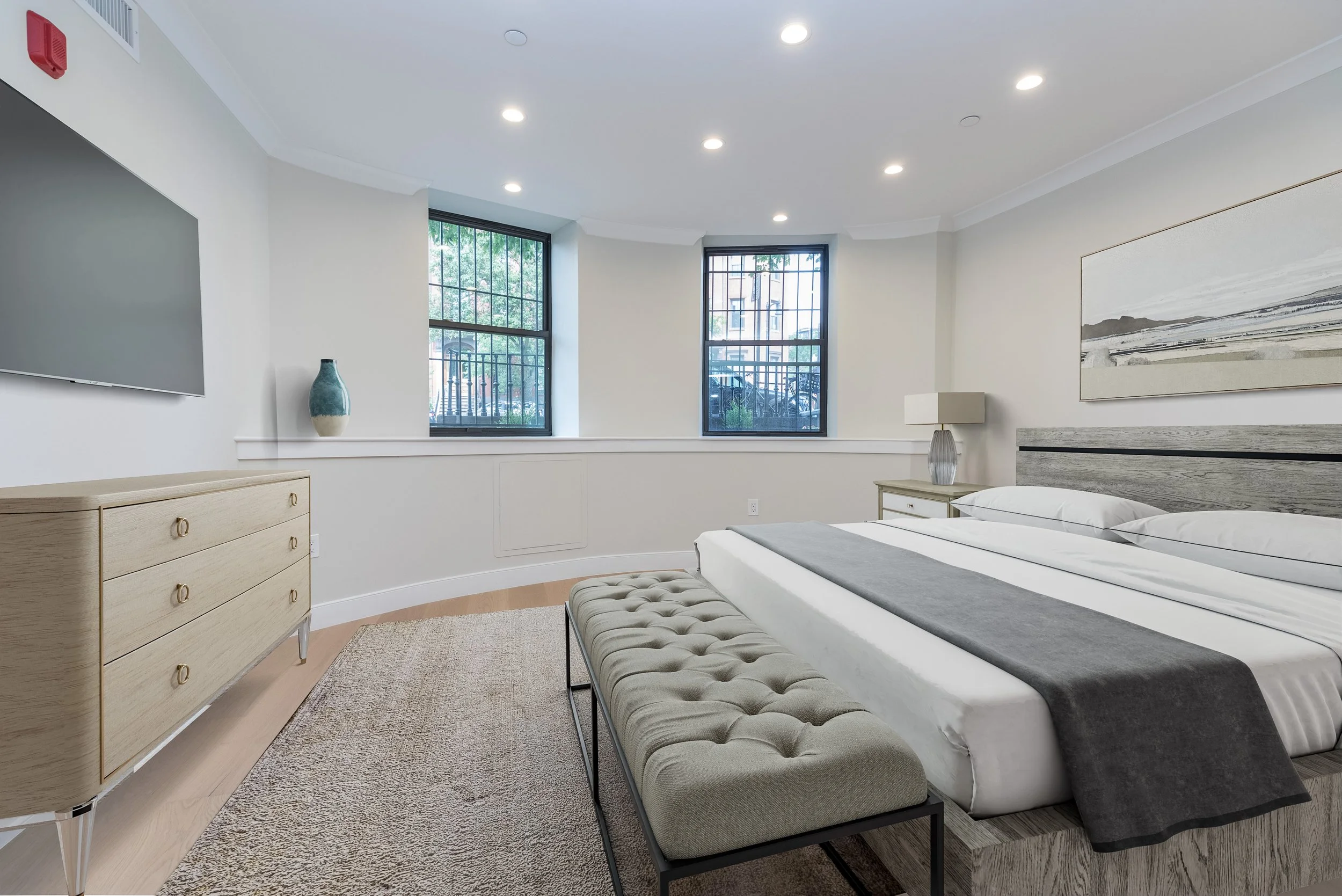 A bedroom with a bed, a dresser, and a side table with a lamp. Two large windows with black frames show an outside view of trees and parked cars. The room has white walls, a beige carpet, and recessed ceiling lights.