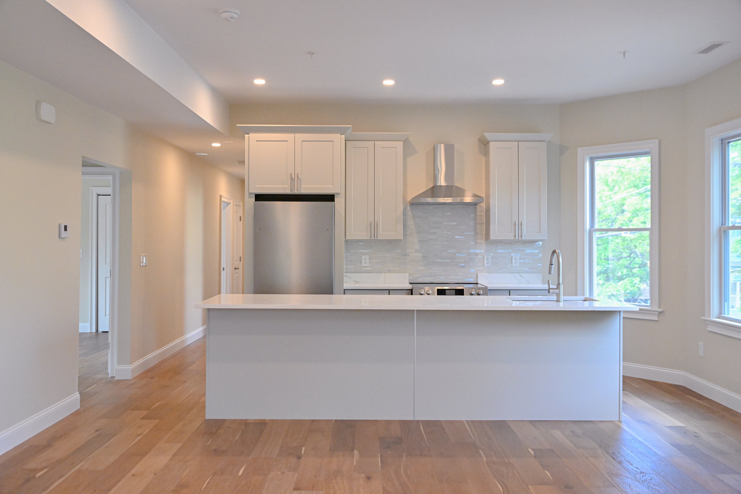 Modern kitchen with white cabinetry and a large kitchen island with a white countertop, stainless steel appliances including a stove and range hood, large windows with views of green trees, and hardwood floors.