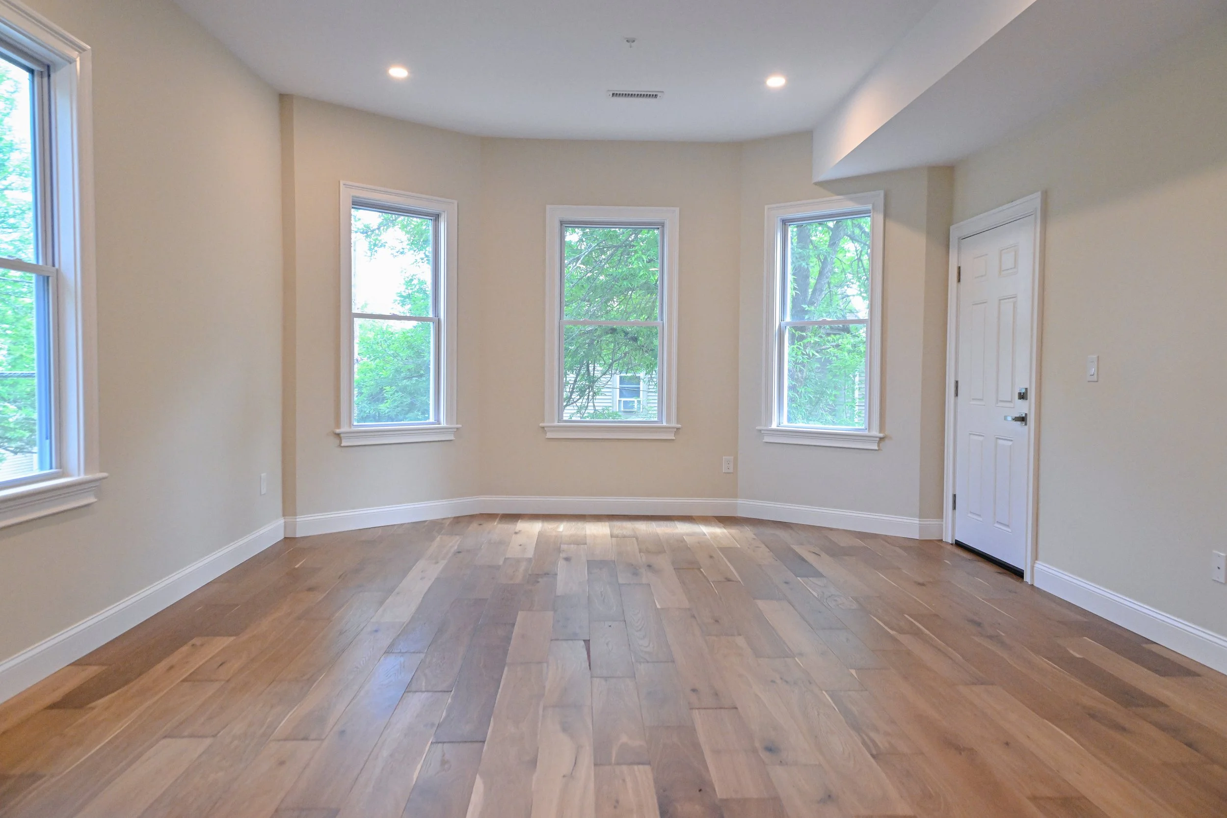 Empty room with hardwood floors, cream-colored walls, large windows with white trim, and a white door.