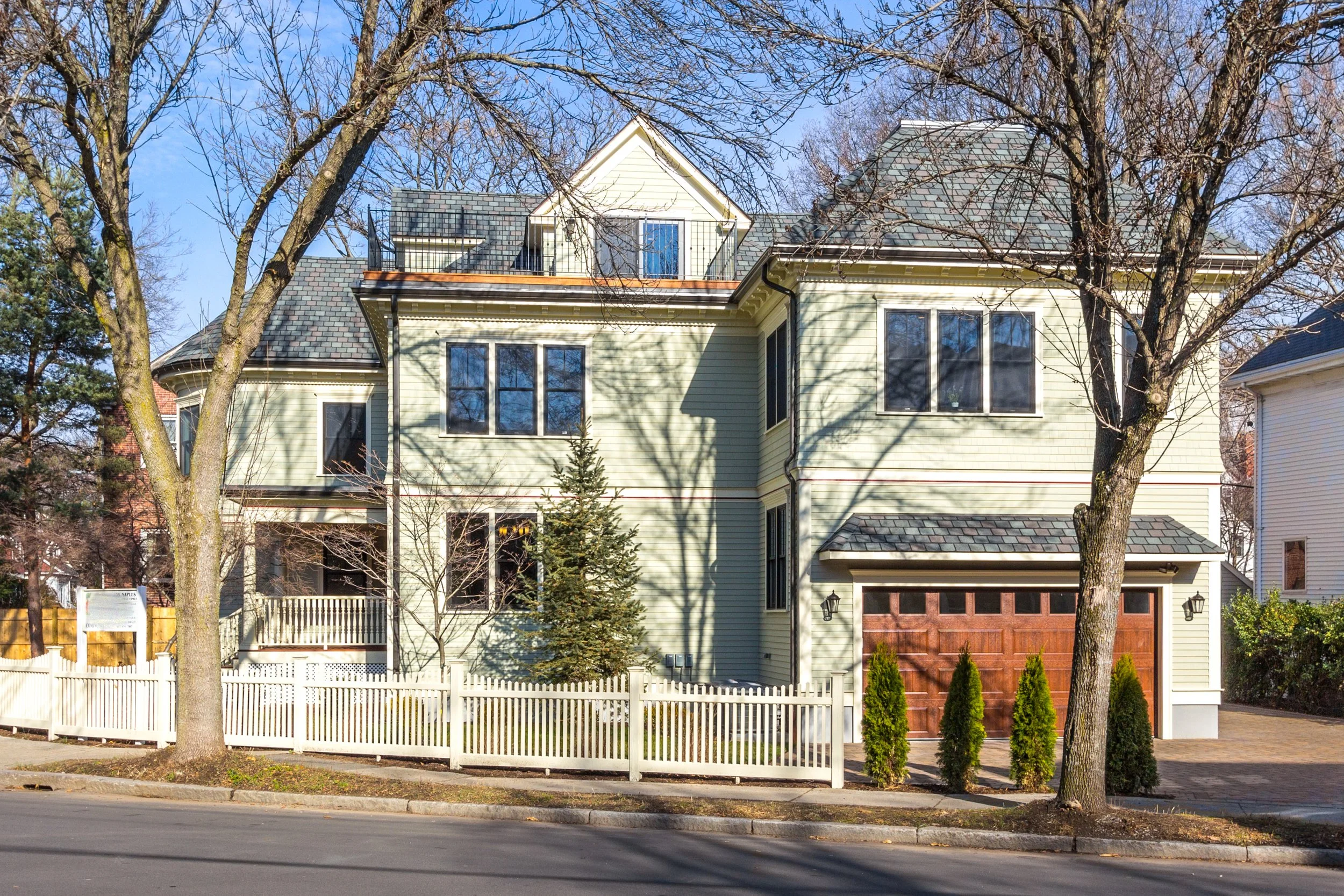 A multi-story yellow house with a gray roof, front porch, and white picket fence, situated on a tree-lined street.