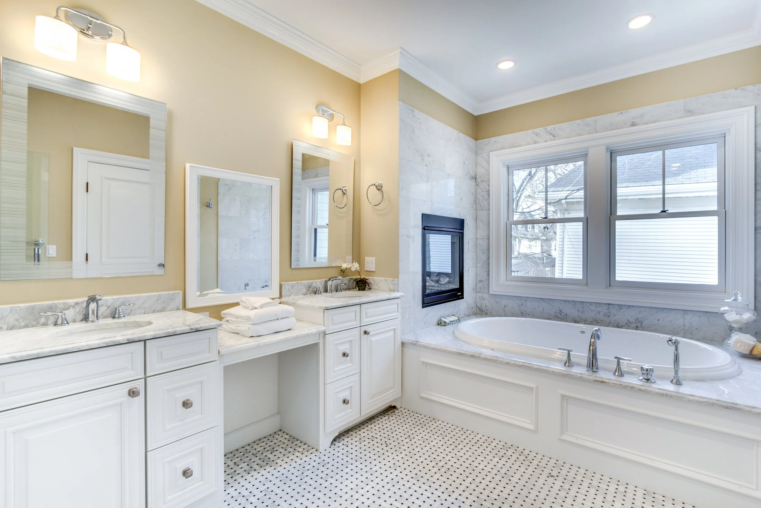Bathroom with double sinks, large mirror, wall-mounted lights, white cabinetry, marble countertops, a bathtub, large window with a view of rooftops, and a marble fireplace.
