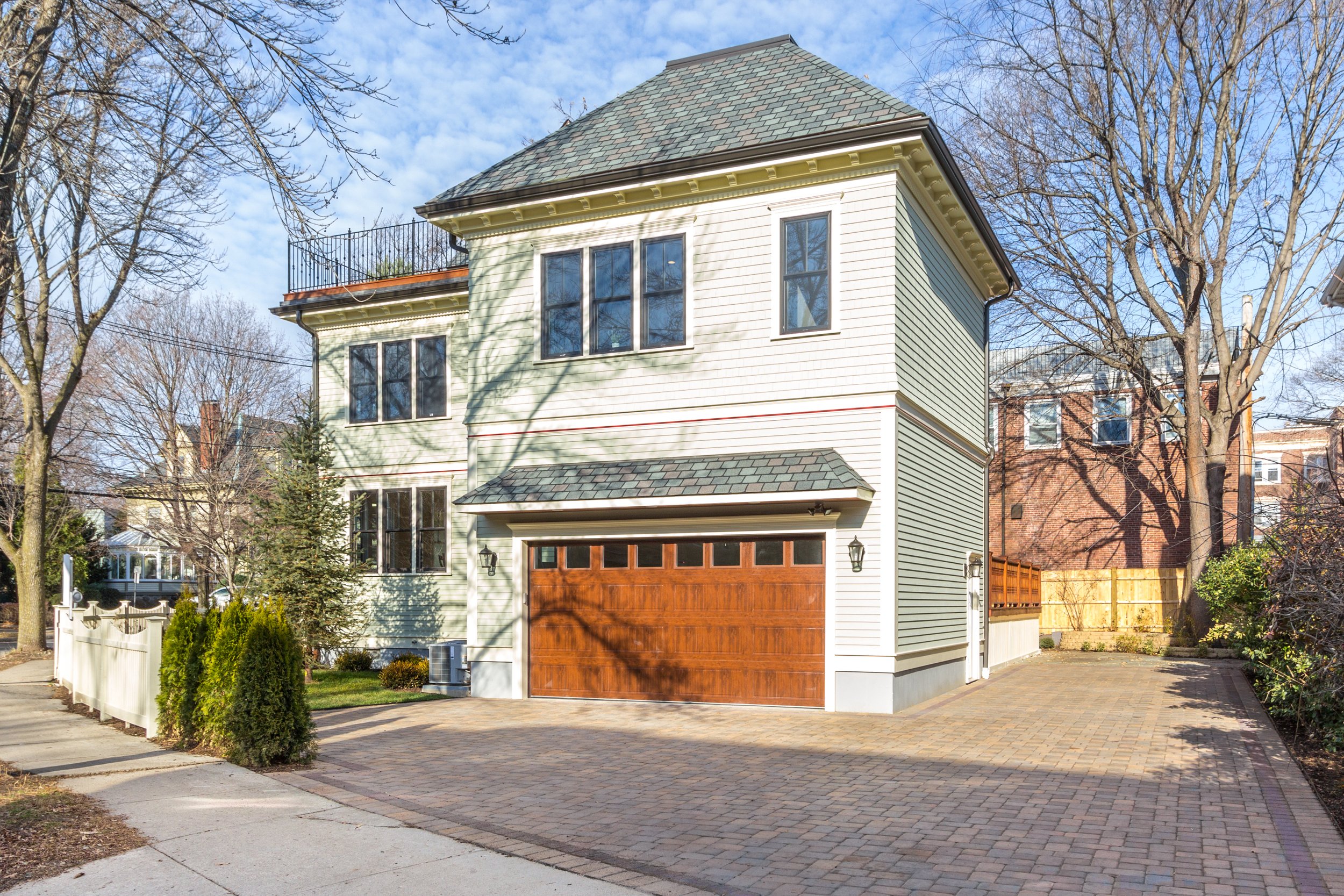 A two-story house with white siding, large windows, a brown garage door, and a gray shingled roof. The house is surrounded by trees with bare branches and a small garden with bushes in the front yard. A brick walkway and driveway are visible.
