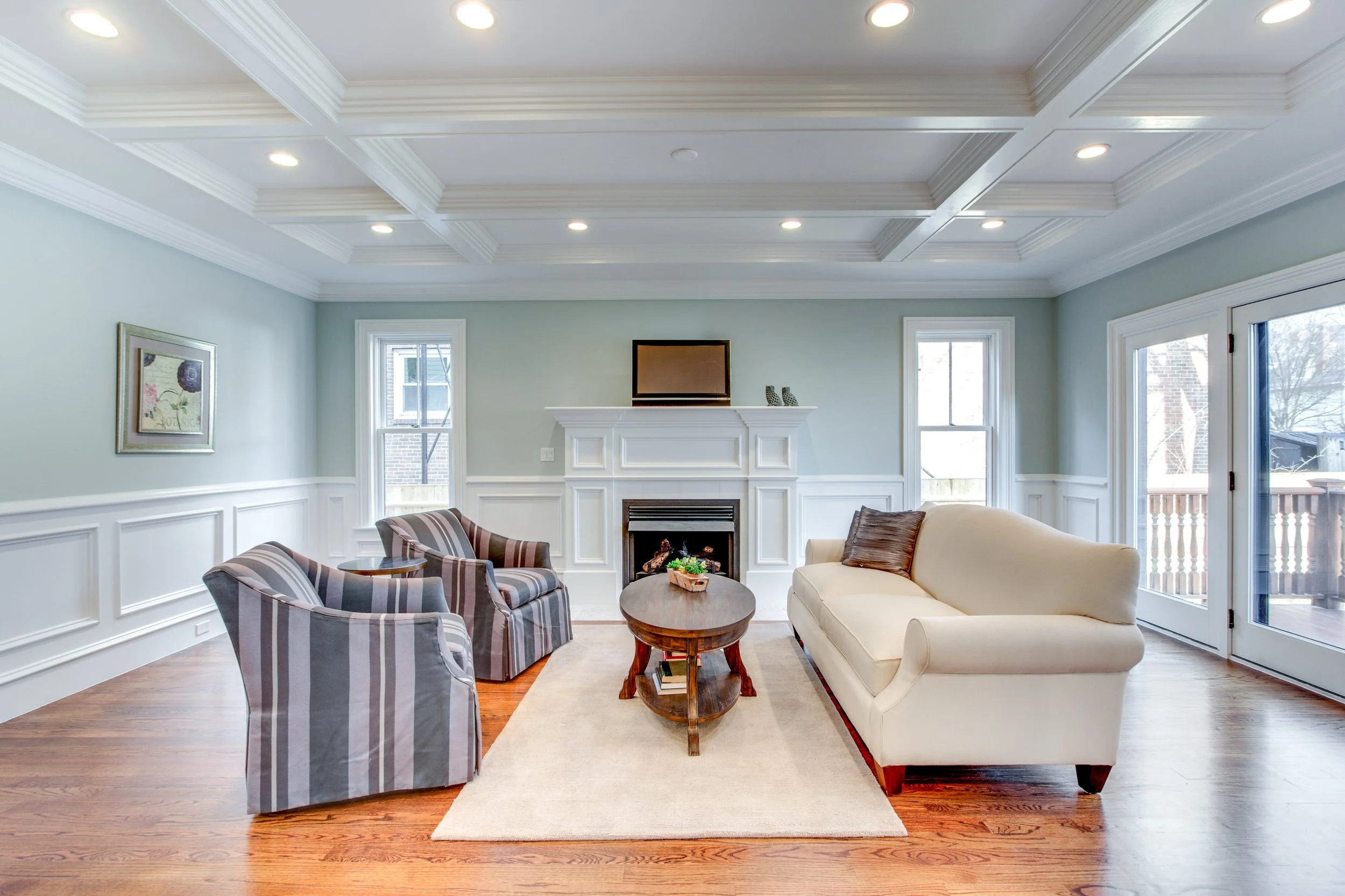 Living room with beige and striped upholstered chairs, a platform fireplace, a beige sofa, a wooden coffee table with a small plant, large windows, and a television on the mantel.