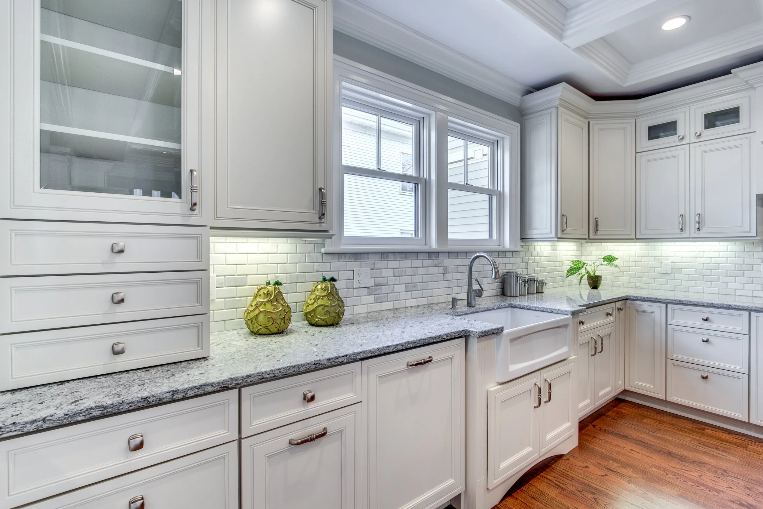 White kitchen with granite countertops, white cabinetry, and a white subway tile backsplash, featuring a sink below three windows and decorative vases on the counter.