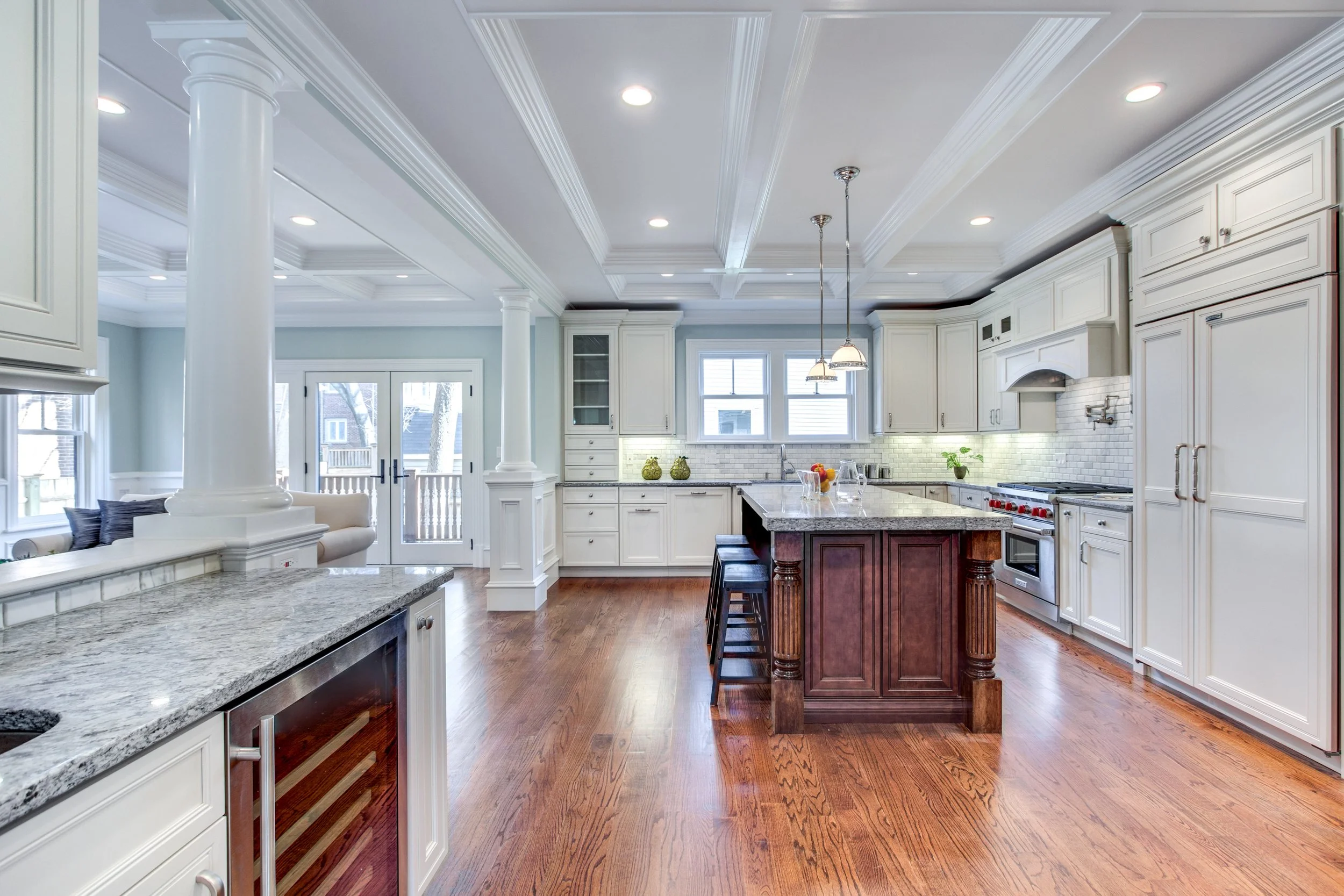 Bright farmhouse kitchen with white cabinets, granite countertops, a central island with dark wood base, hardwood floors, and large windows bringing in natural light.