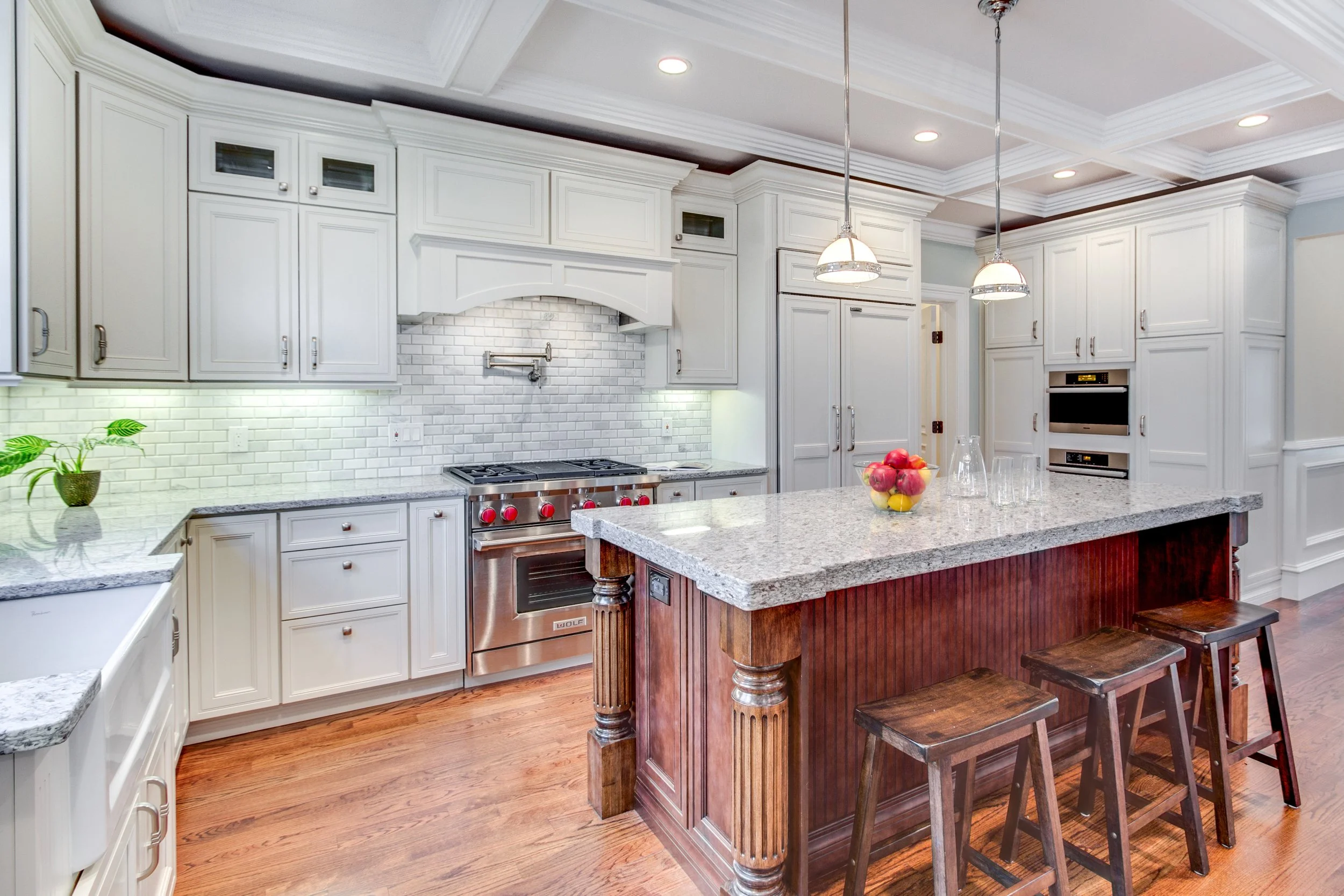 Modern kitchen with white cabinets, granite countertops, stainless steel oven, and a large island with wooden stools, pendant lights, fruit bowl, and hardwood flooring.