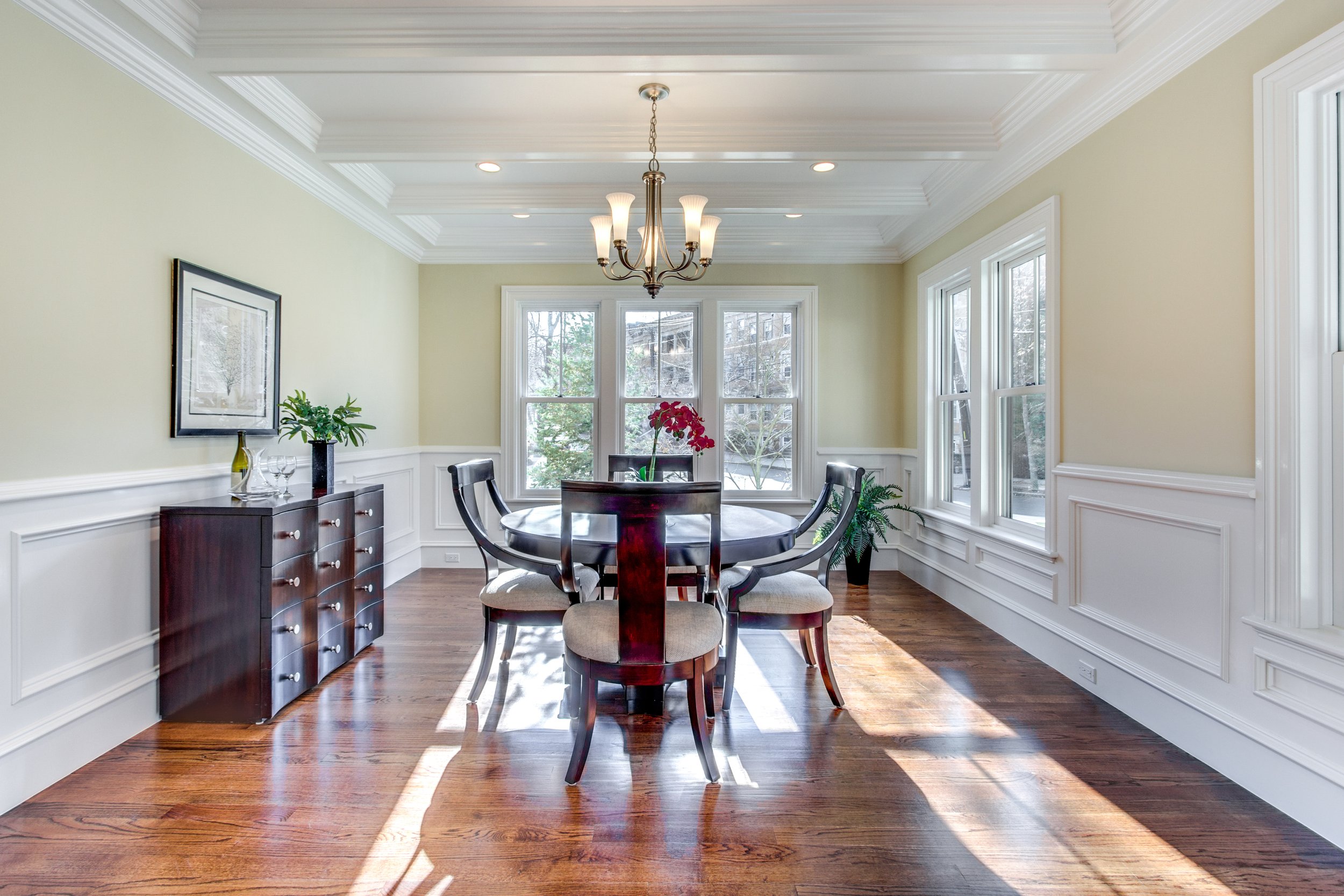 Dining room with round table, six chairs, chandelier, large windows, hardwood floor, and wall decor.