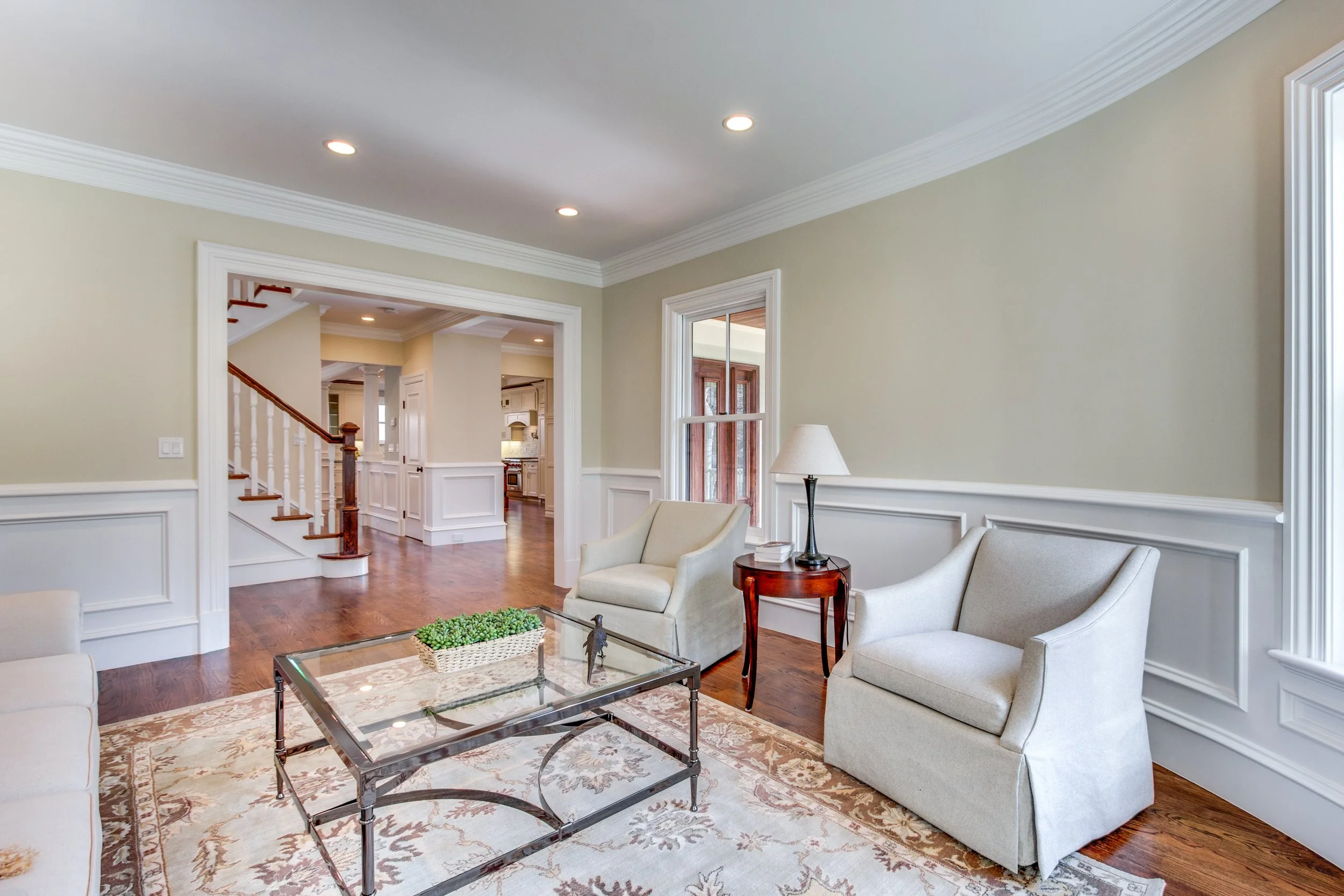 Living room with cream-colored walls, white crown molding, hardwood floors, and large windows. Furniture includes a beige armchair, a cream sofa, a glass coffee table with a metal frame, a small wooden side table with a lamp, and a decorative plant. 