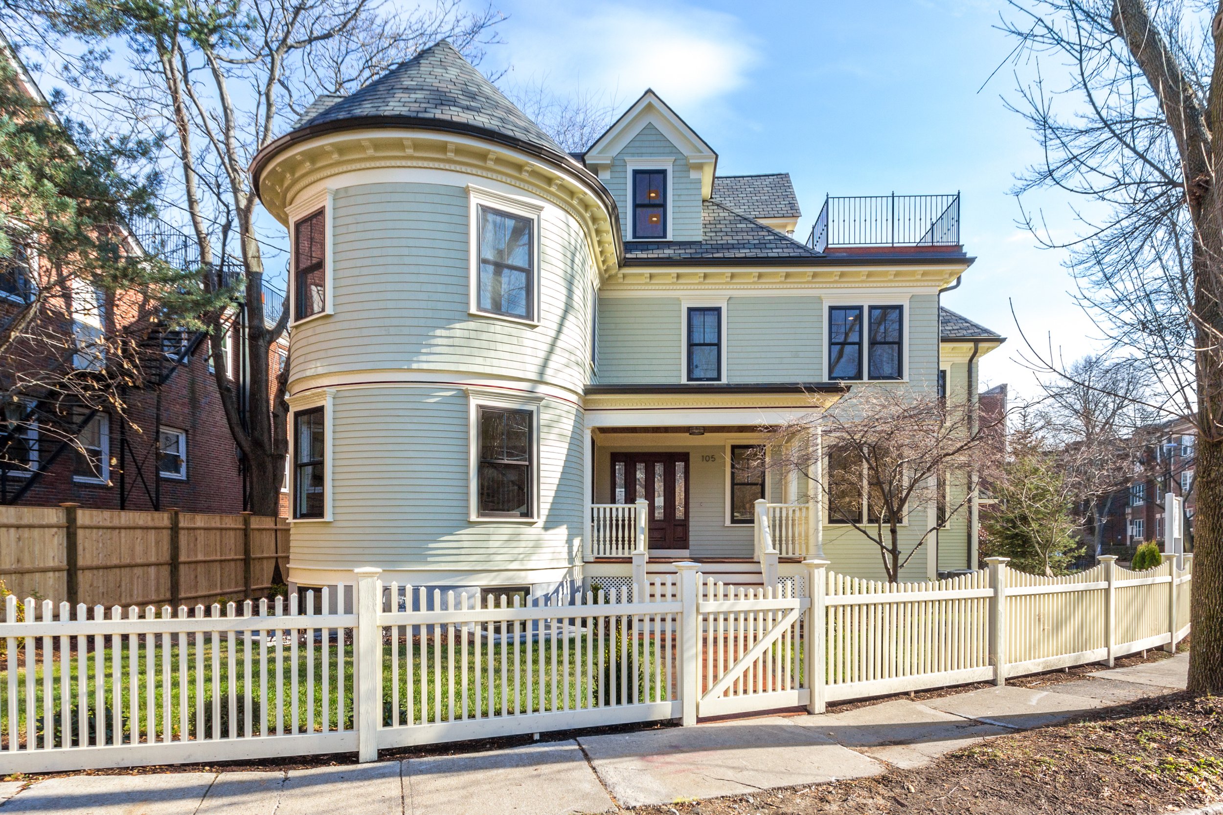 A Victorian-style house with pale yellow siding, a prominent rounded turret with windows, a gabled roof, and a front porch with white railing. The house is surrounded by trees and a white picket fence, with a sidewalk in front.
