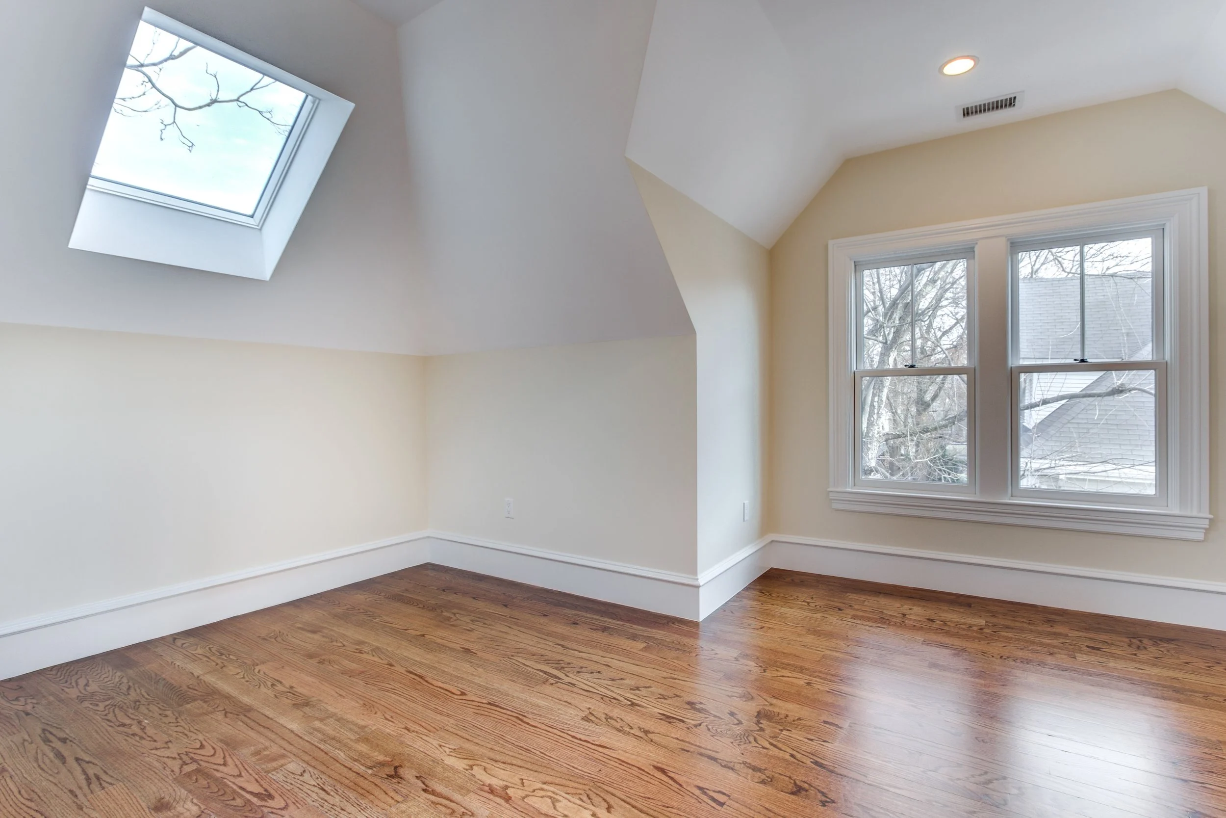 A room with hardwood flooring, two windows on the right wall, and a skylight window on the ceiling. The walls are painted light cream, and the ceiling has recessed lighting. Outside the windows, trees can be seen.
