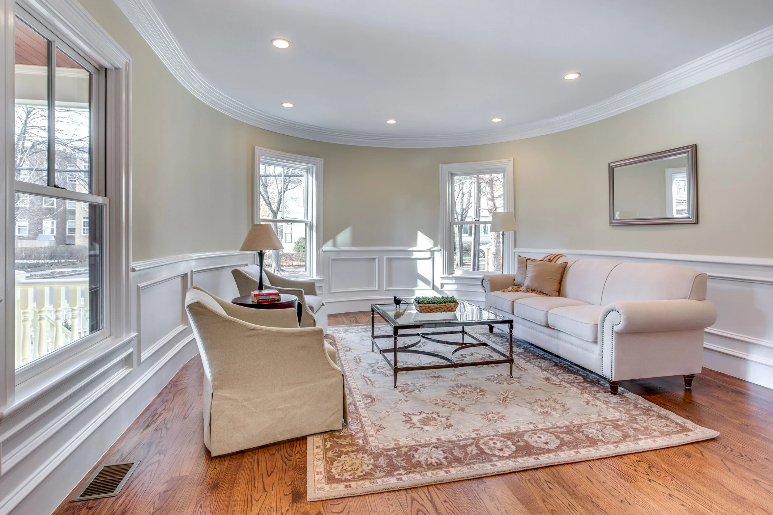 Living room with beige sofa, armchairs, glass coffee table, area rug, and multiple windows showing outside trees, with ceiling lights and crown molding.