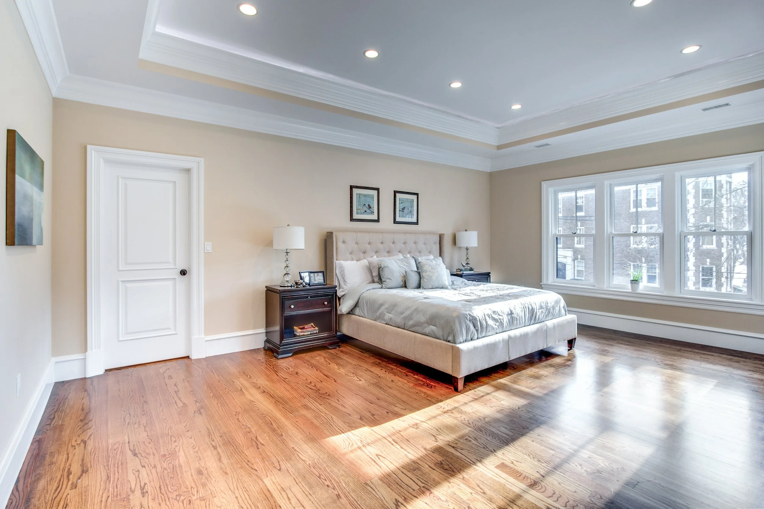 A bedroom with wooden floor, beige walls, a bed with a tufted beige headboard, two nightstands with lamps, and large windows letting in natural light.