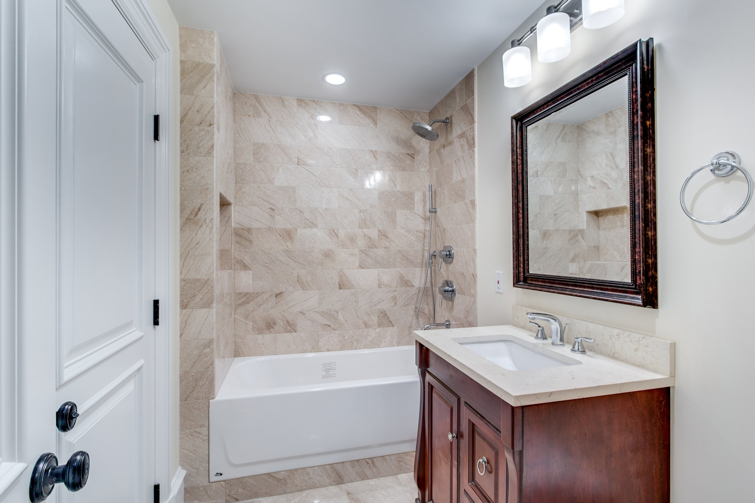 Bathroom with beige tiled shower, white bathtub, wooden vanity with sink, large mirror, and wall-mounted light fixtures.
