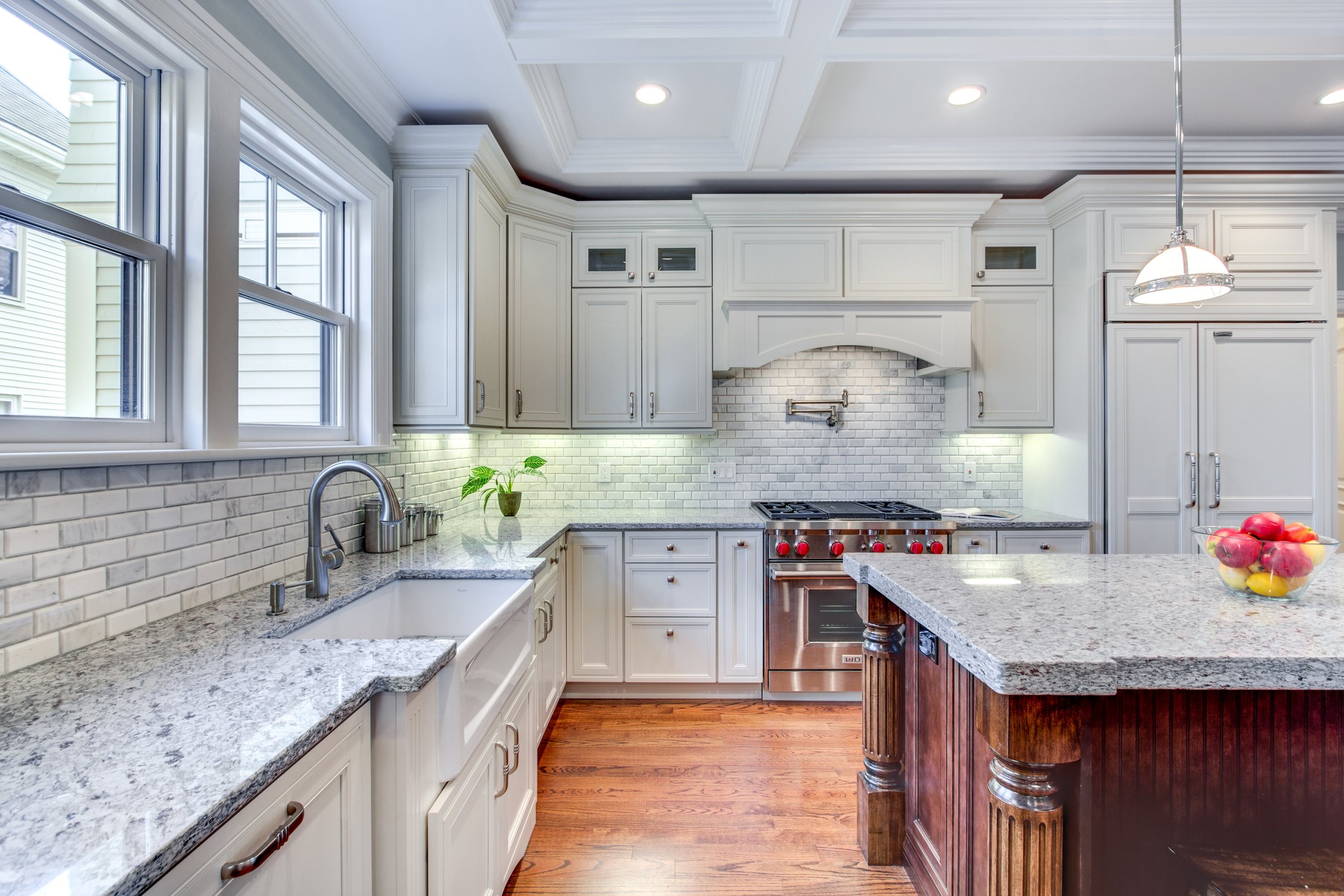 A modern kitchen with large windows, white cabinets, granite countertops, a gas stove, and a kitchen island with a bowl of apples.