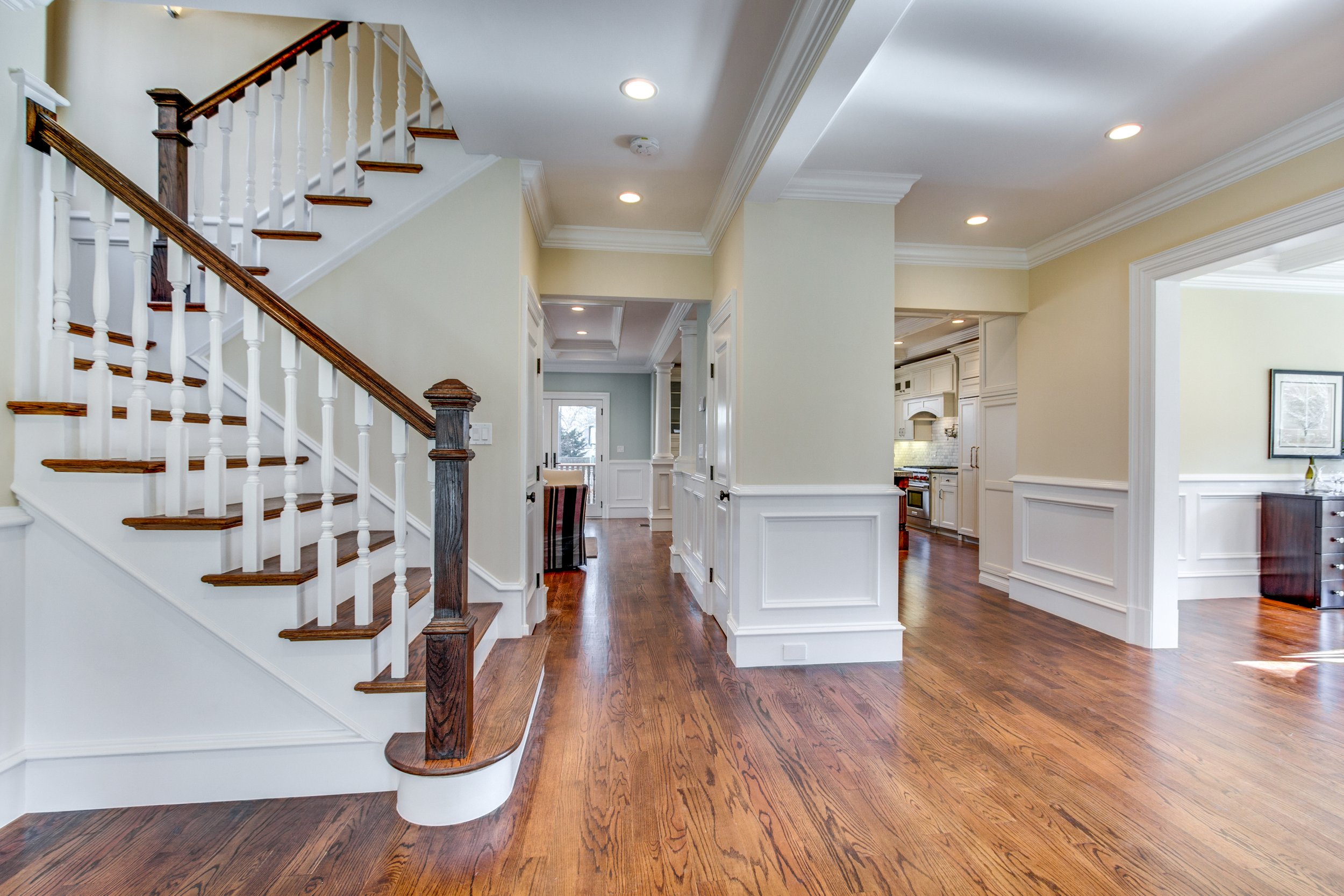 Interior view of a spacious home with hardwood floors, white and beige walls, a staircase with wooden handrails, and a kitchen area with white cabinetry.