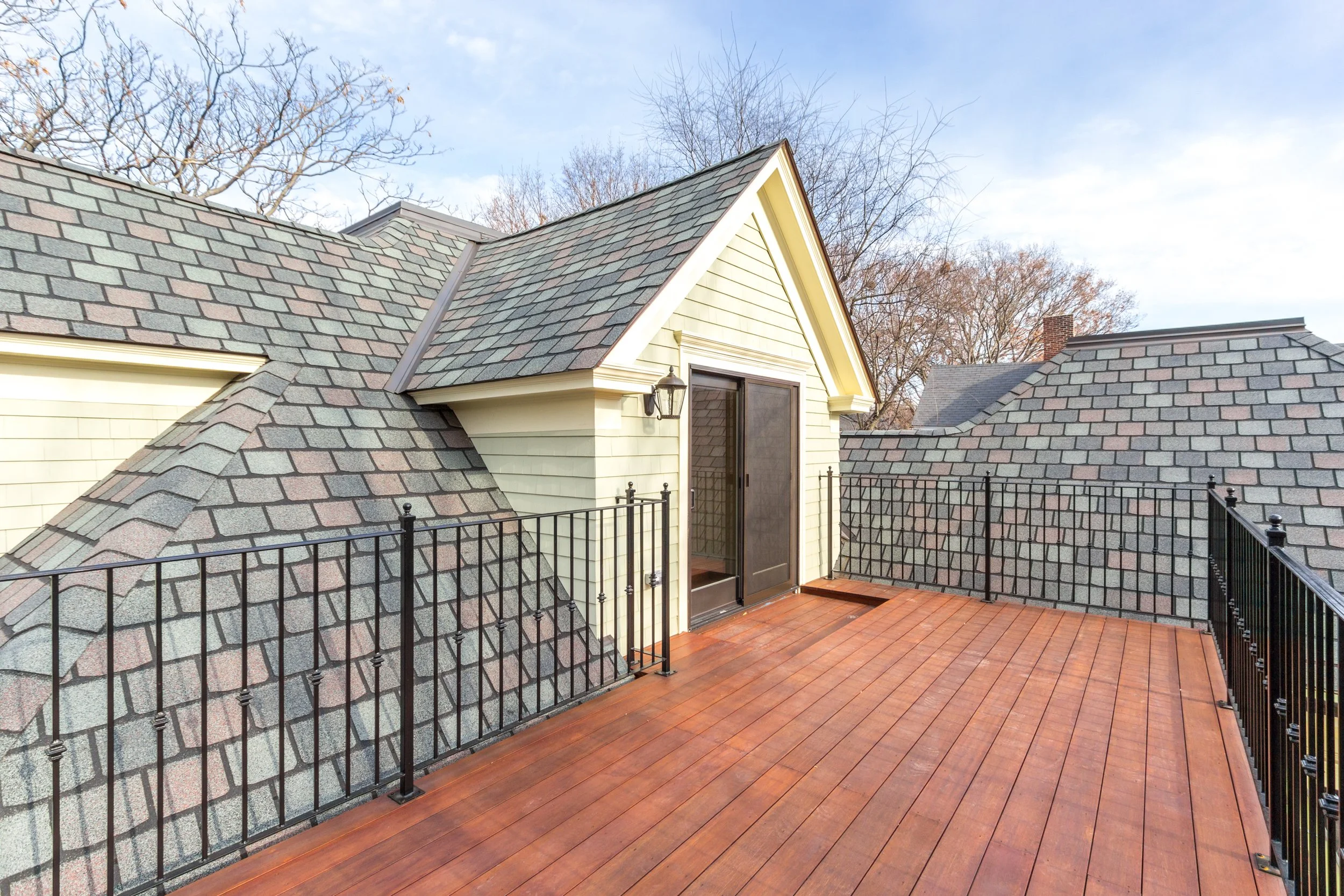 A rooftop balcony with wooden flooring, black metal railing, beige house siding, and a door. The roof has gray shingles, and there are leafless trees and a chimney in the background against a blue sky.