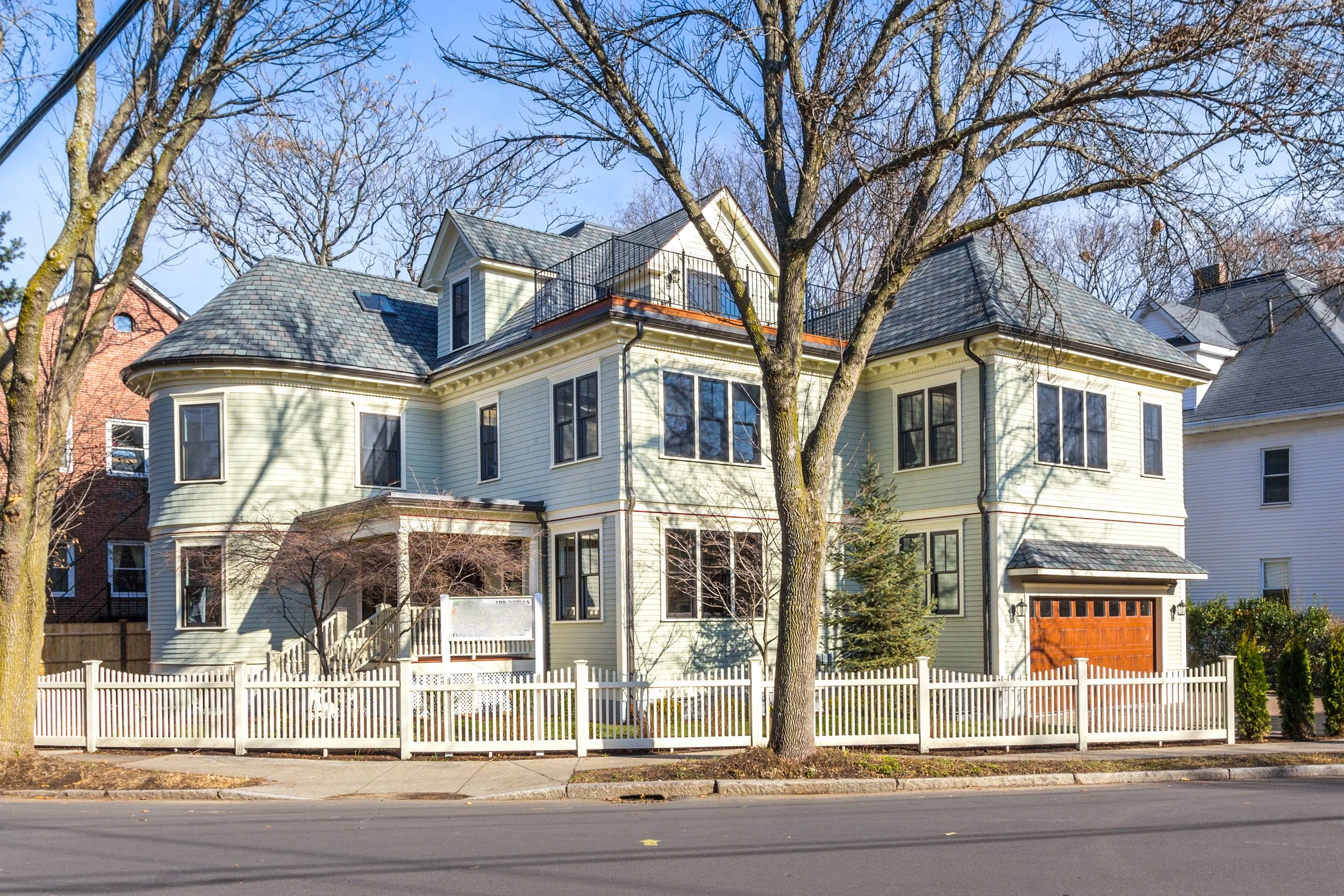 A light yellow multi-story house with a white picket fence, several leafless trees in front, and neighboring houses visible in the background under a clear blue sky.