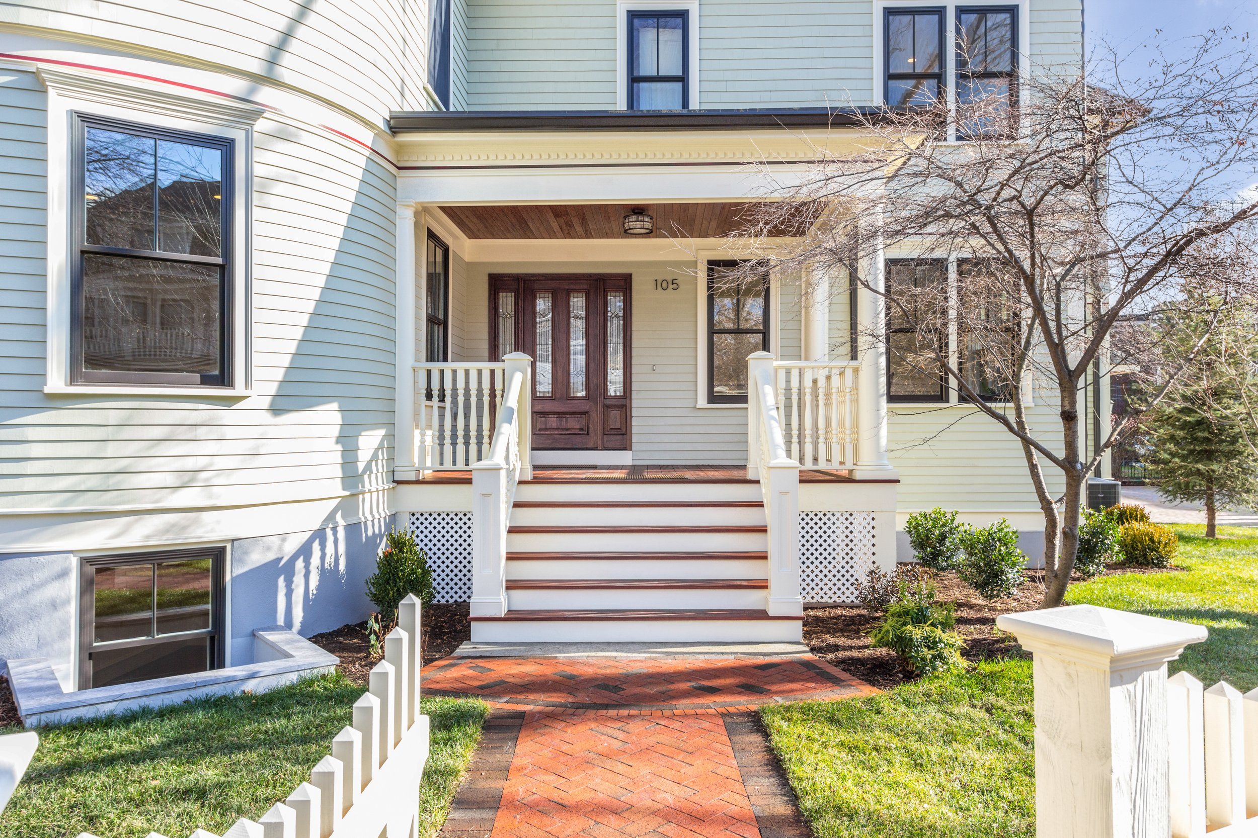 Front porch of a yellow house with white trim, a brown front door, stairs, and a red brick walkway. There is a small tree with bare branches and shrubs in front.