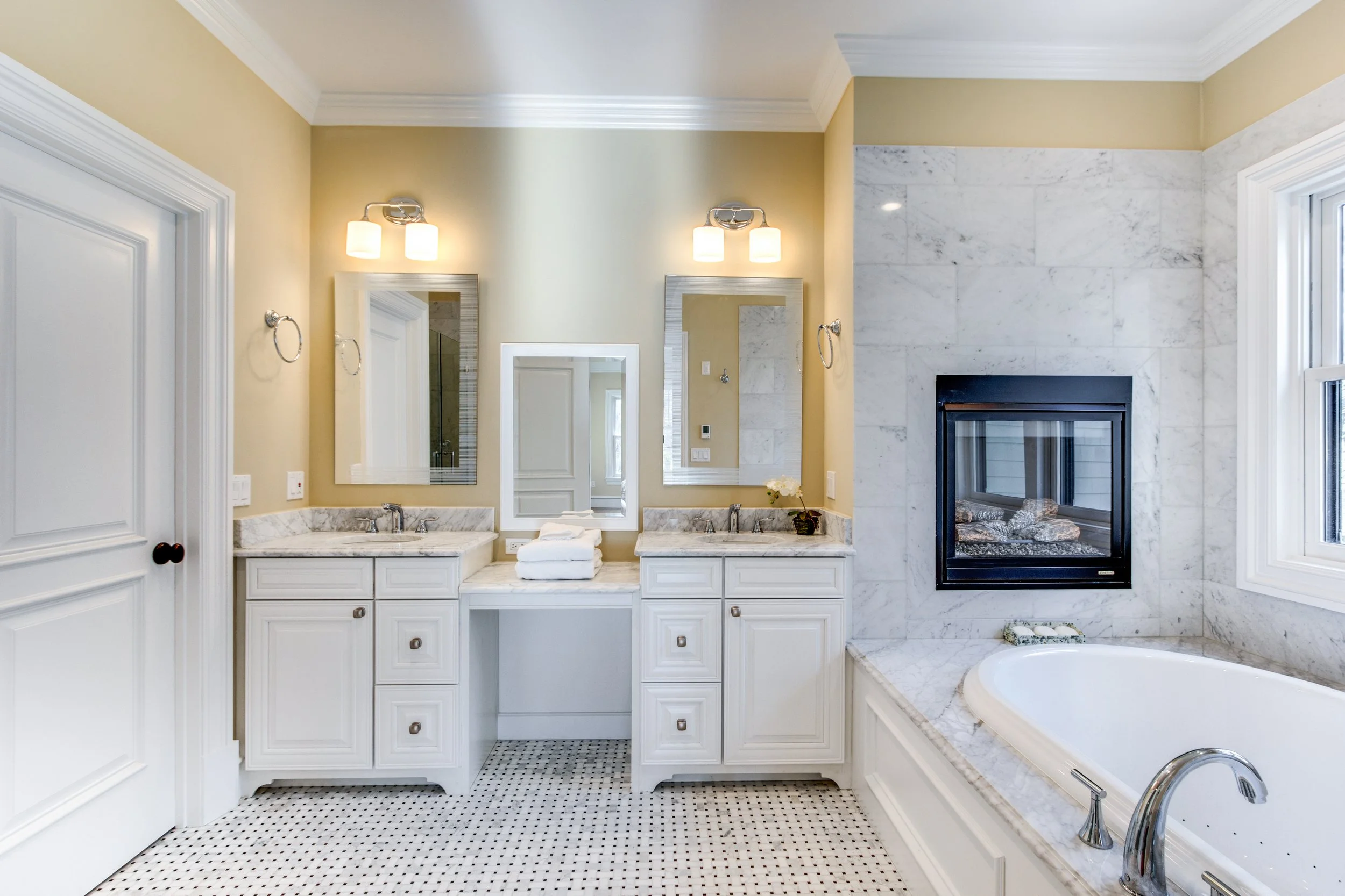 Bathroom with double sink vanity, mirrors, wall sconces, a fireplace with marble surround, and a bathtub with marble surround.