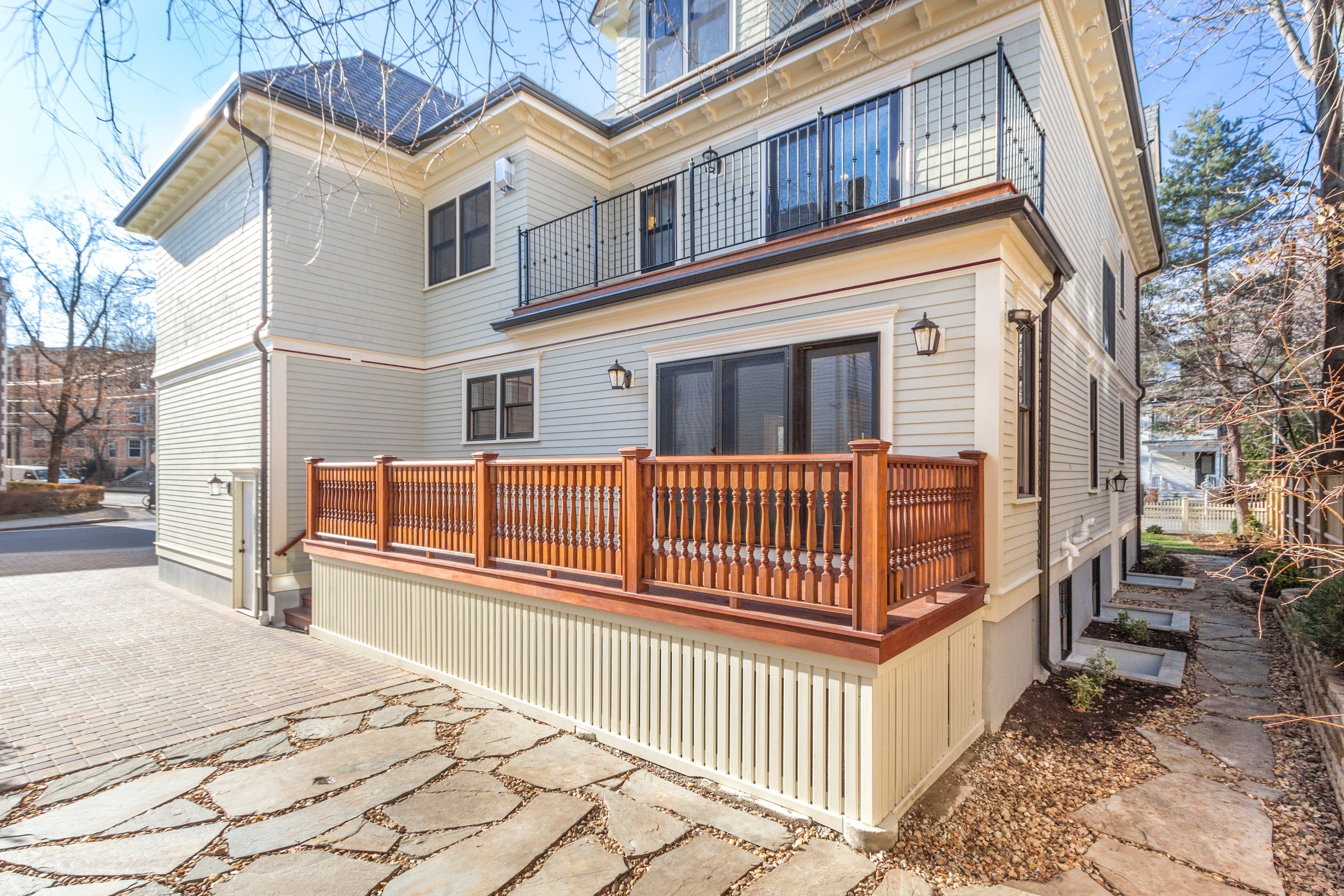 A modern two-story house with beige siding, black window frames, and a rooftop balcony with black metal railings. The house has outdoor lighting fixtures and a small wooden deck extending from the lower level, enclosed by a wooden railing.