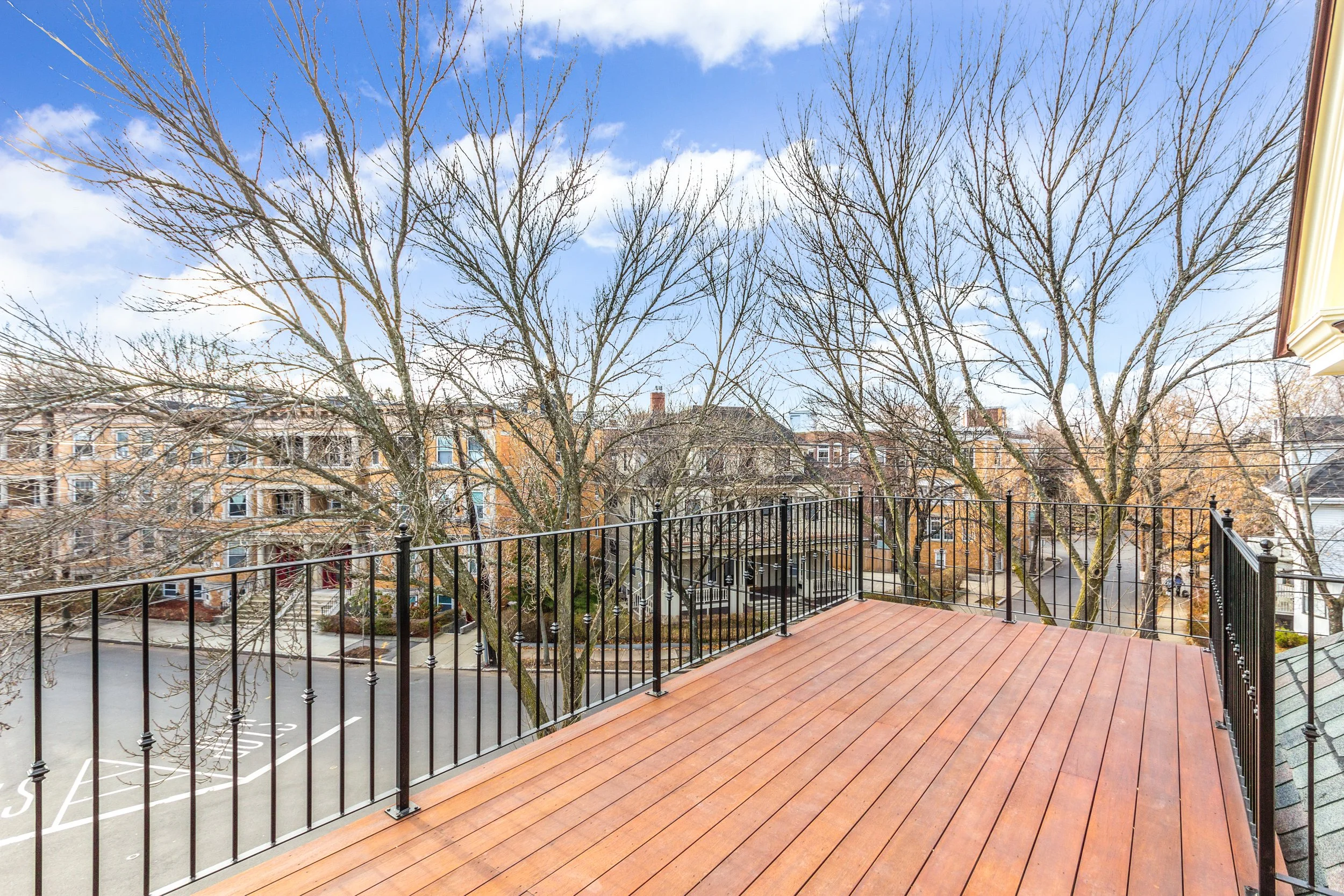 View from a balcony with a black metal railing and wooden floorboards, overlooking residential buildings and leafless trees in a neighborhood under a partly cloudy blue sky.
