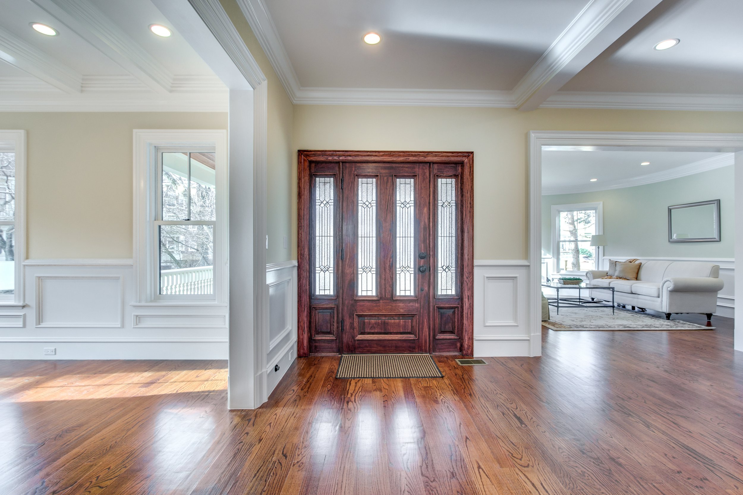 Interior of a house with wooden floors, cream walls, crown molding, and a red front door with decorative glass panels, leading into a living room with a sofa, coffee table, and windows.