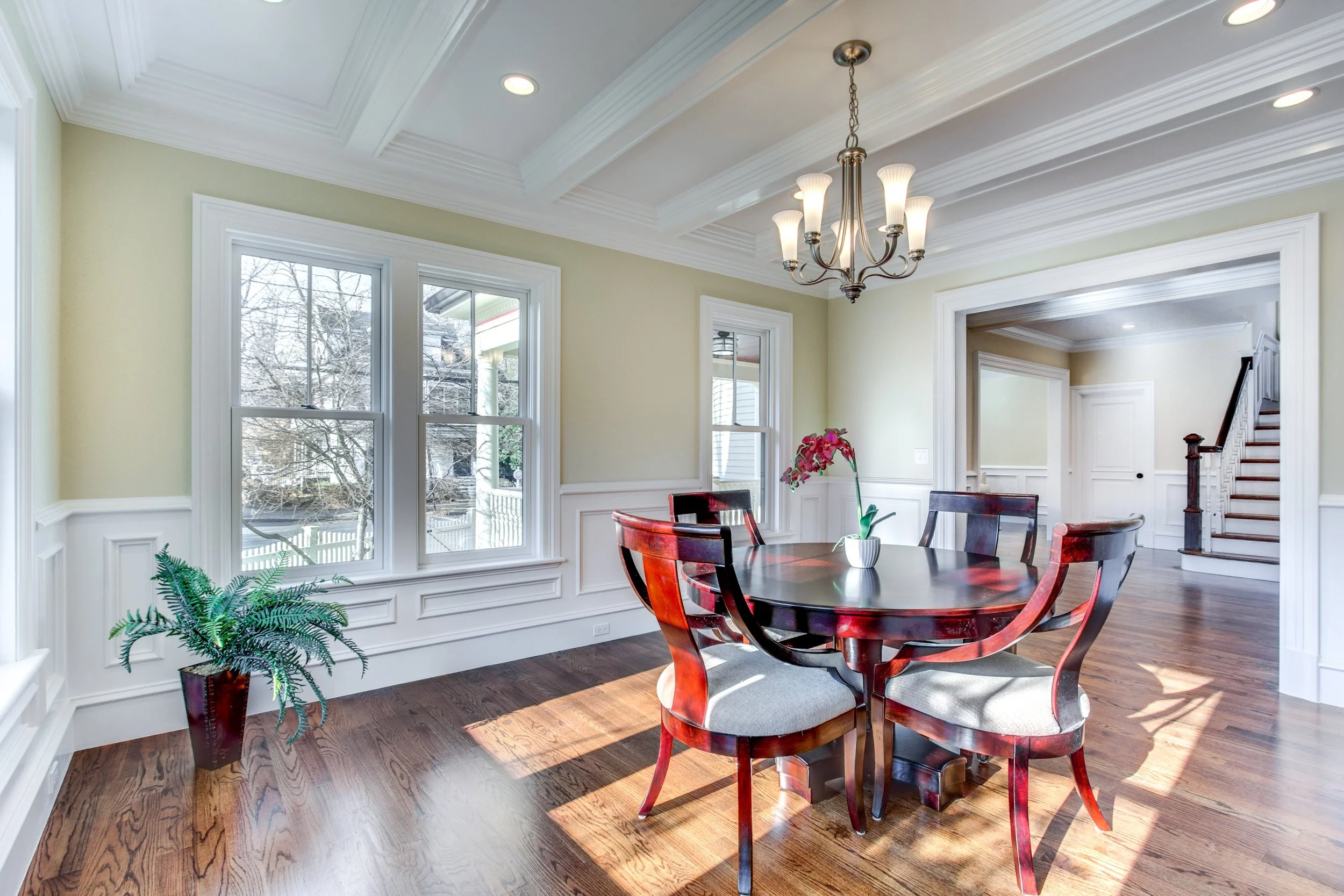 A dining room with large windows, a round wooden table, and six matching chairs. There is a flower arrangement on the table and a potted plant on the floor. The ceiling has recessed lighting and a chandelier, with hardwood floors and wainscoting on t