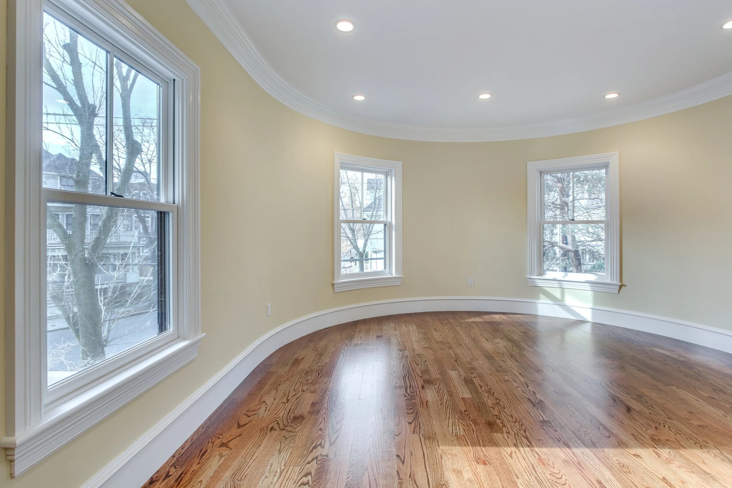 Empty room with three large windows, hardwood floor, yellow walls and white crown molding, during daytime.