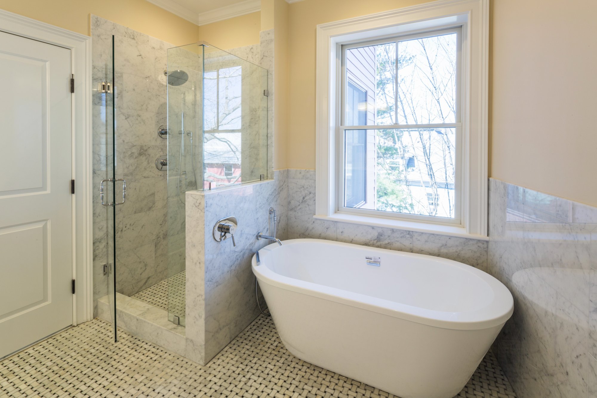 A modern bathroom with a glass-enclosed shower, a freestanding bathtub, a large window with white trim, and marble tilework.