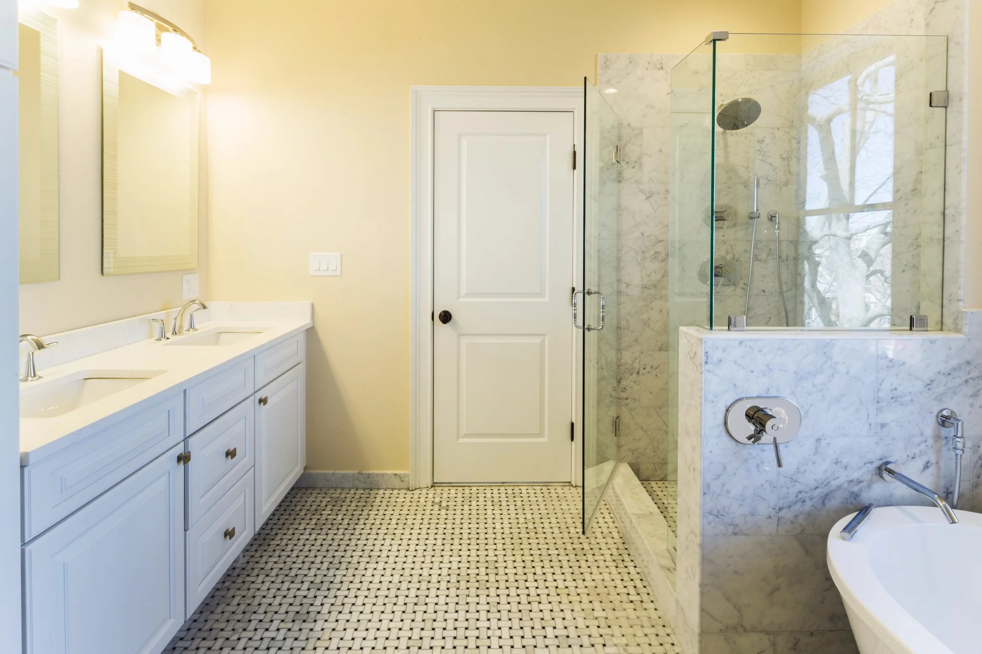 A clean bathroom with a double sink vanity, a glass-enclosed shower, and a white door. The floor has small black and white mosaic tiles, and the walls are painted a soft yellow. There are two large mirrors above the vanity, with lighting fixtures abo