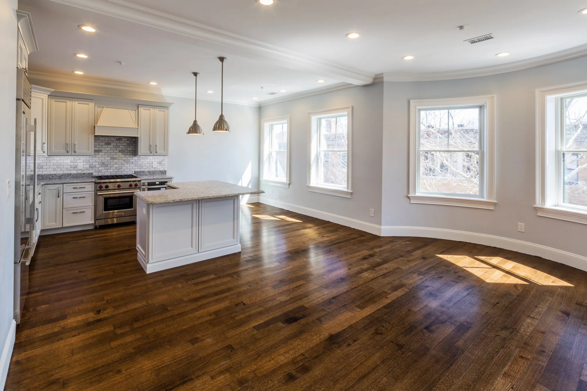 Bright, spacious kitchen with white cabinets, granite countertops, a stainless steel oven, and multiple large windows allowing natural light to fill the room. Hardwood floors are visible throughout.