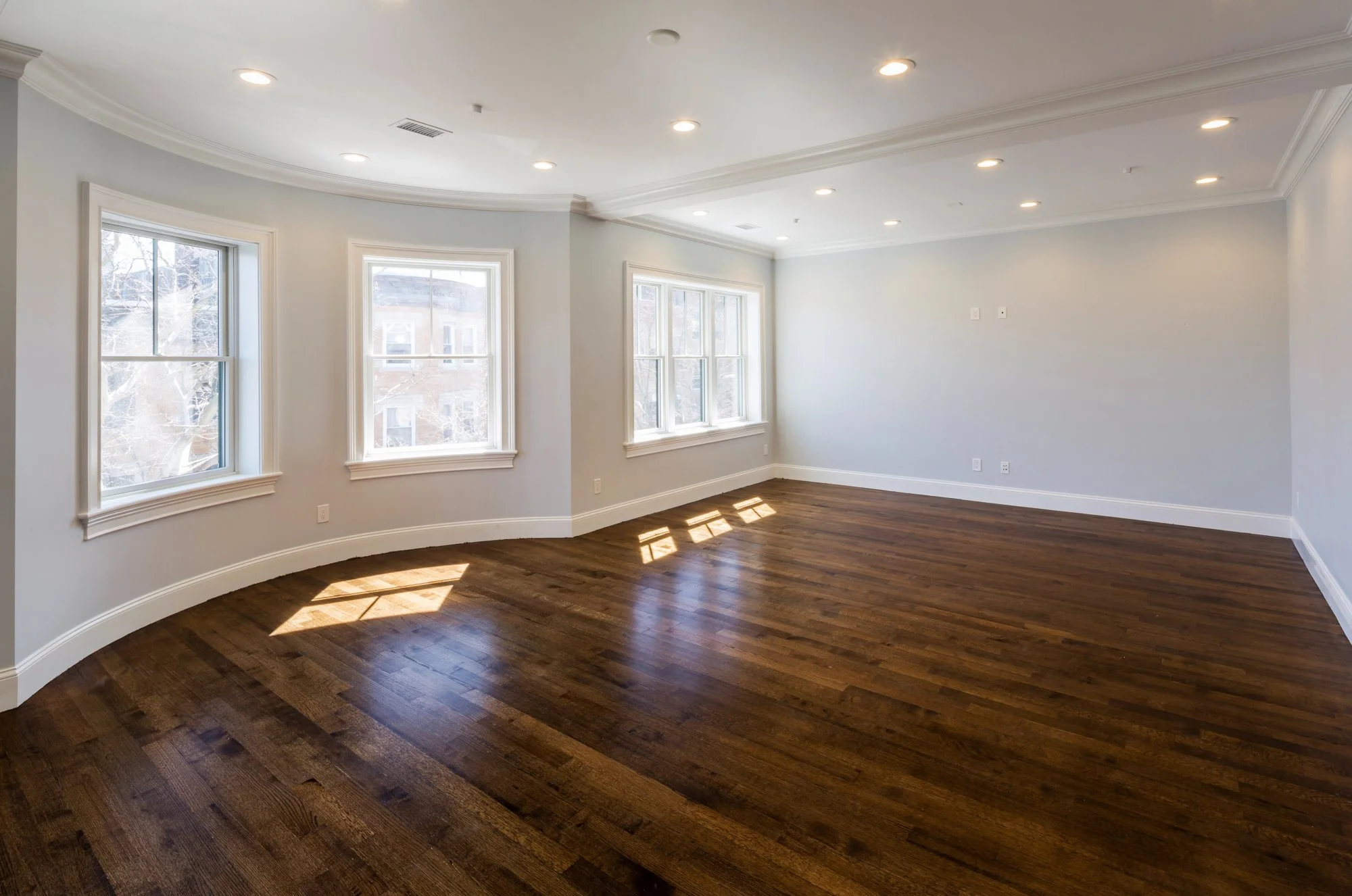 Empty living room with hardwood floors, white walls, large windows, crown molding, and recessed lighting.