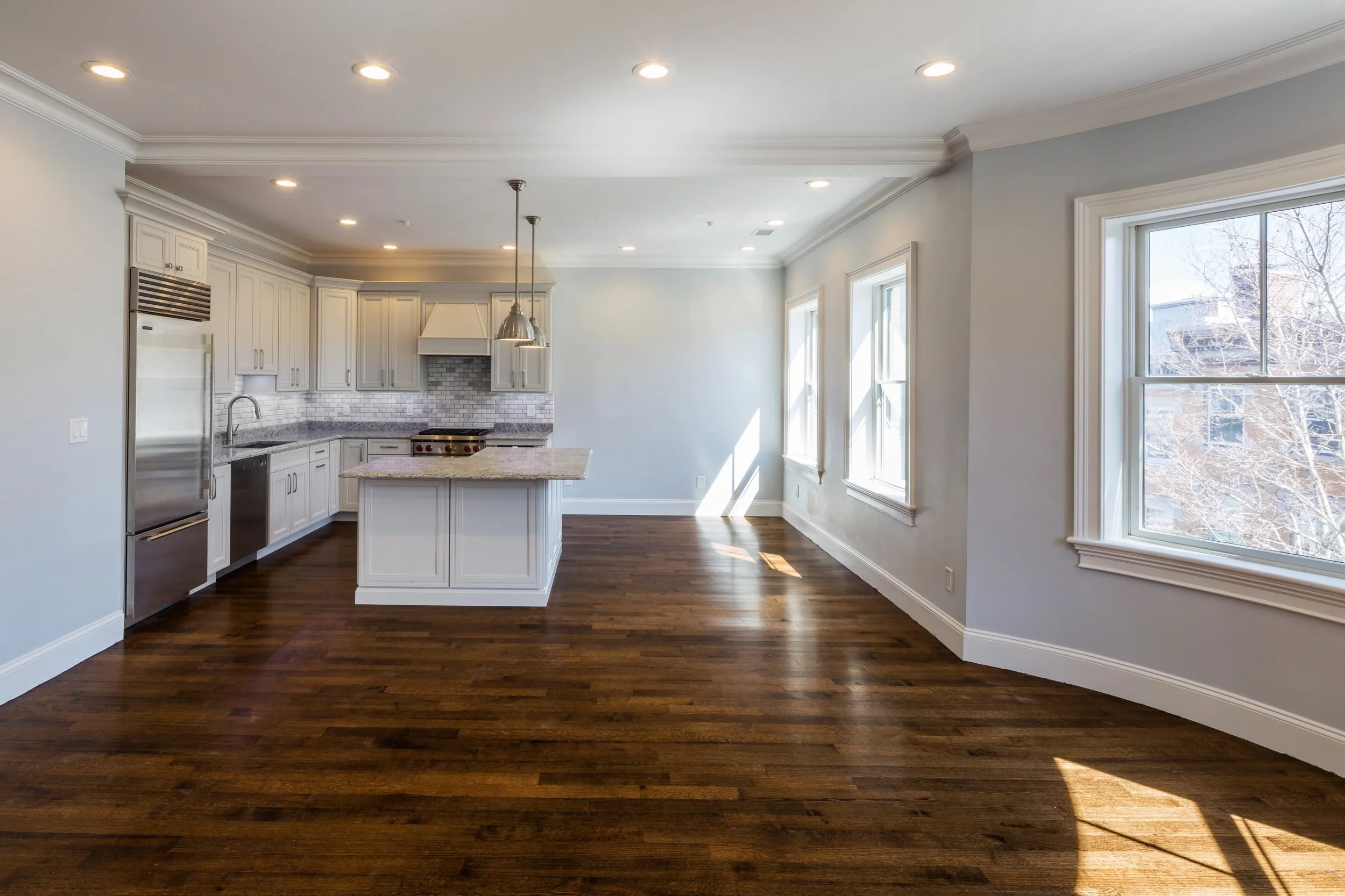 Empty kitchen with white cabinets, granite countertops, and dark hardwood floors, illuminated by recessed ceiling lights and natural light through multiple large windows.