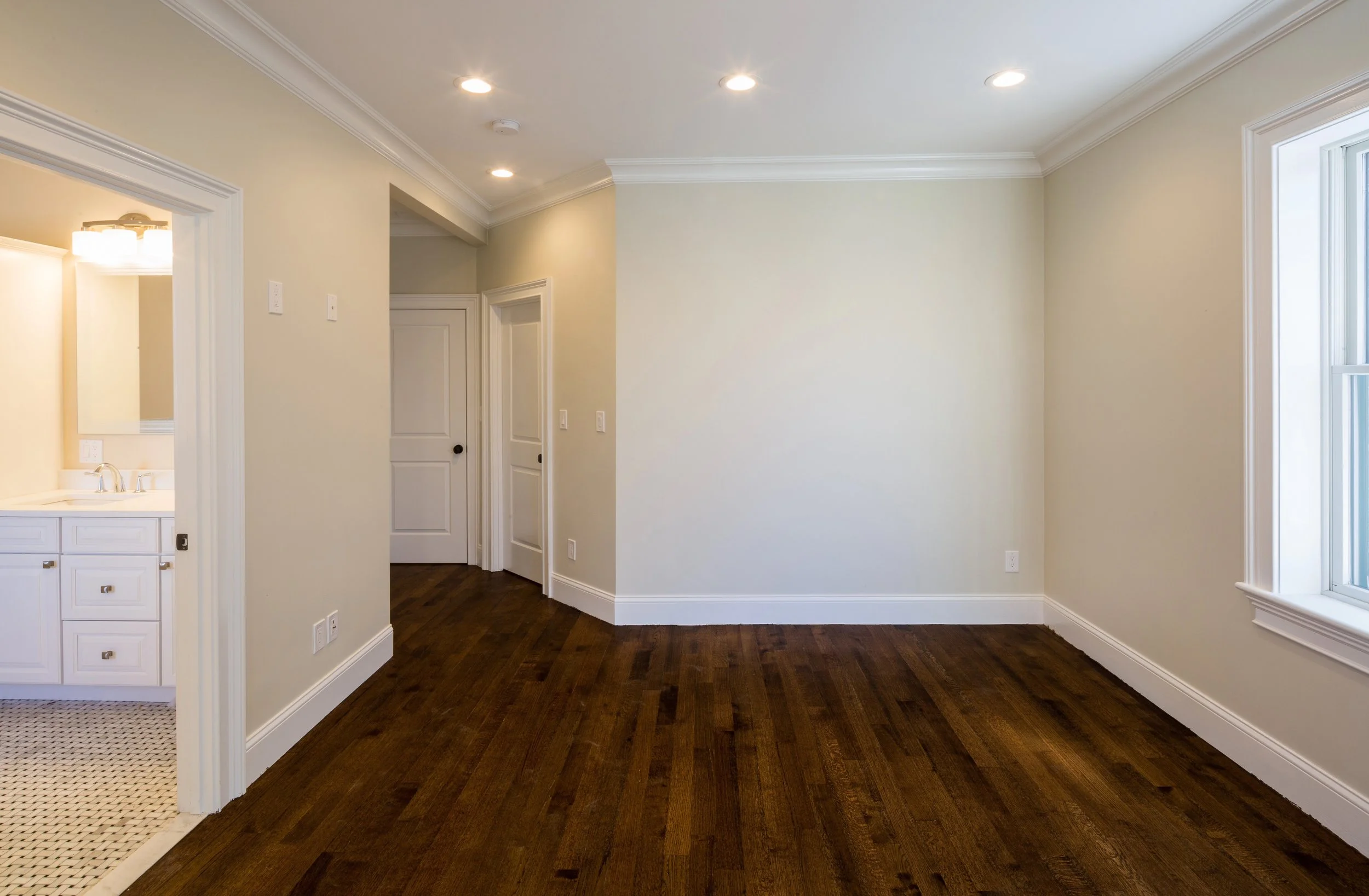 Empty room with hardwood floors, cream-colored walls, white crown molding, and a large window on the right. Recessed lighting on the ceiling and a view into a bathroom with white cabinetry on the left.