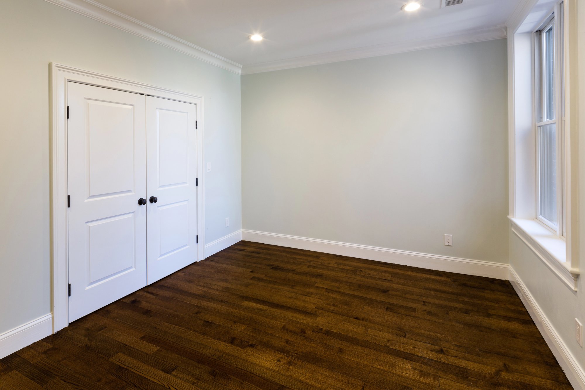 Empty room with hardwood flooring, white walls, a double closet with white doors, a window with white trim, and recessed ceiling lights.