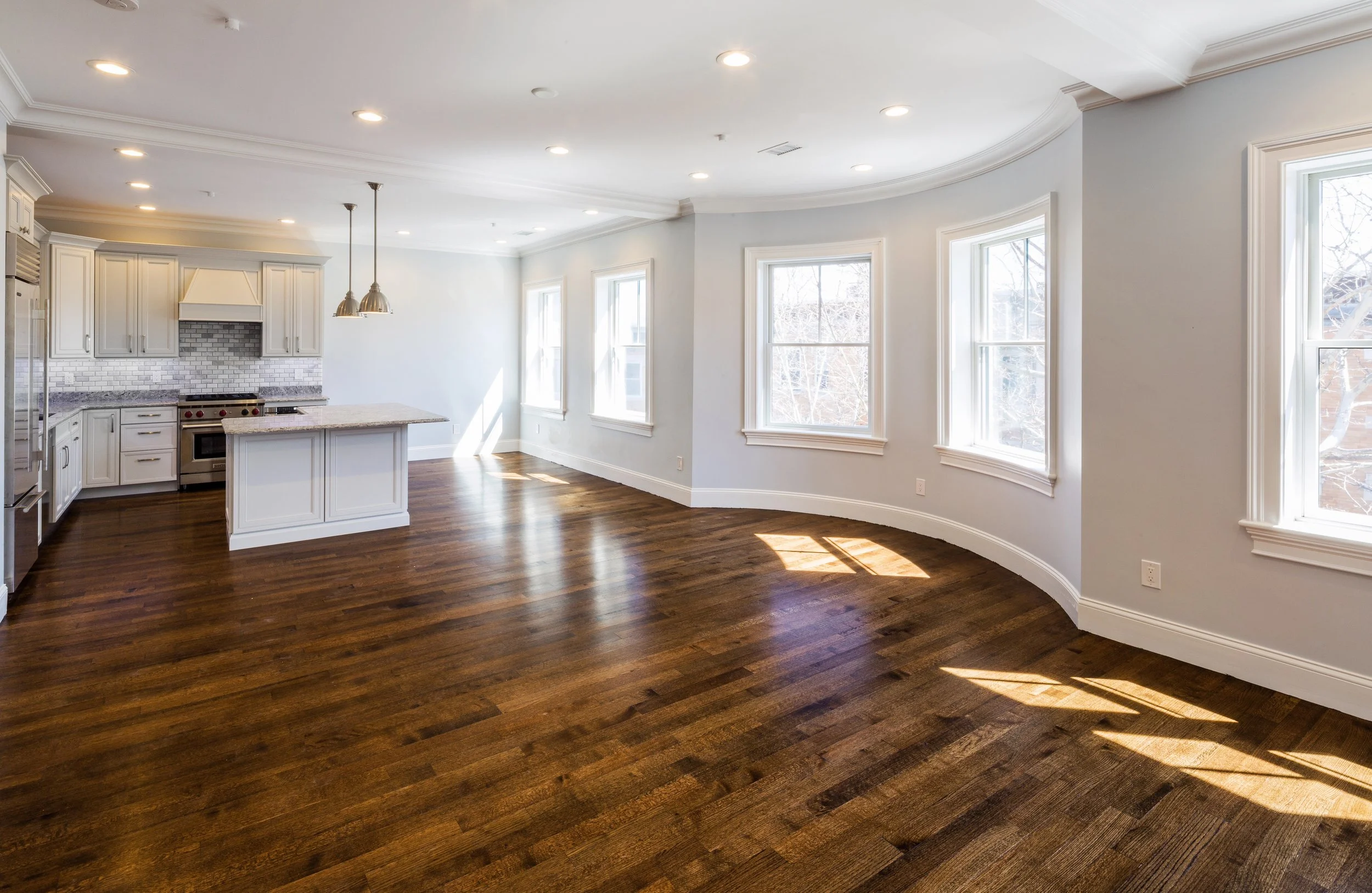 Empty modern kitchen with white cabinets, granite countertops, hardwood floors, and multiple large windows letting in natural light.