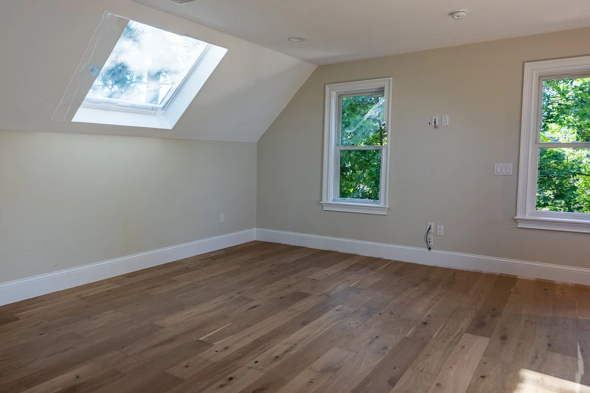 Empty room with hardwood floors, beige walls, white baseboards, two windows with view of trees, and a skylight in the sloped ceiling.