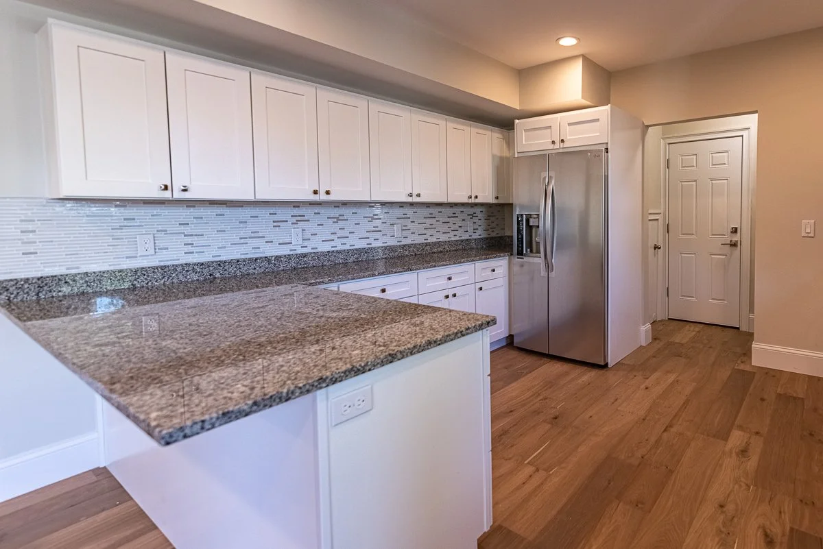 Modern kitchen with white cabinets, stainless steel refrigerator, granite countertops, mosaic tile backsplash, and wooden flooring.