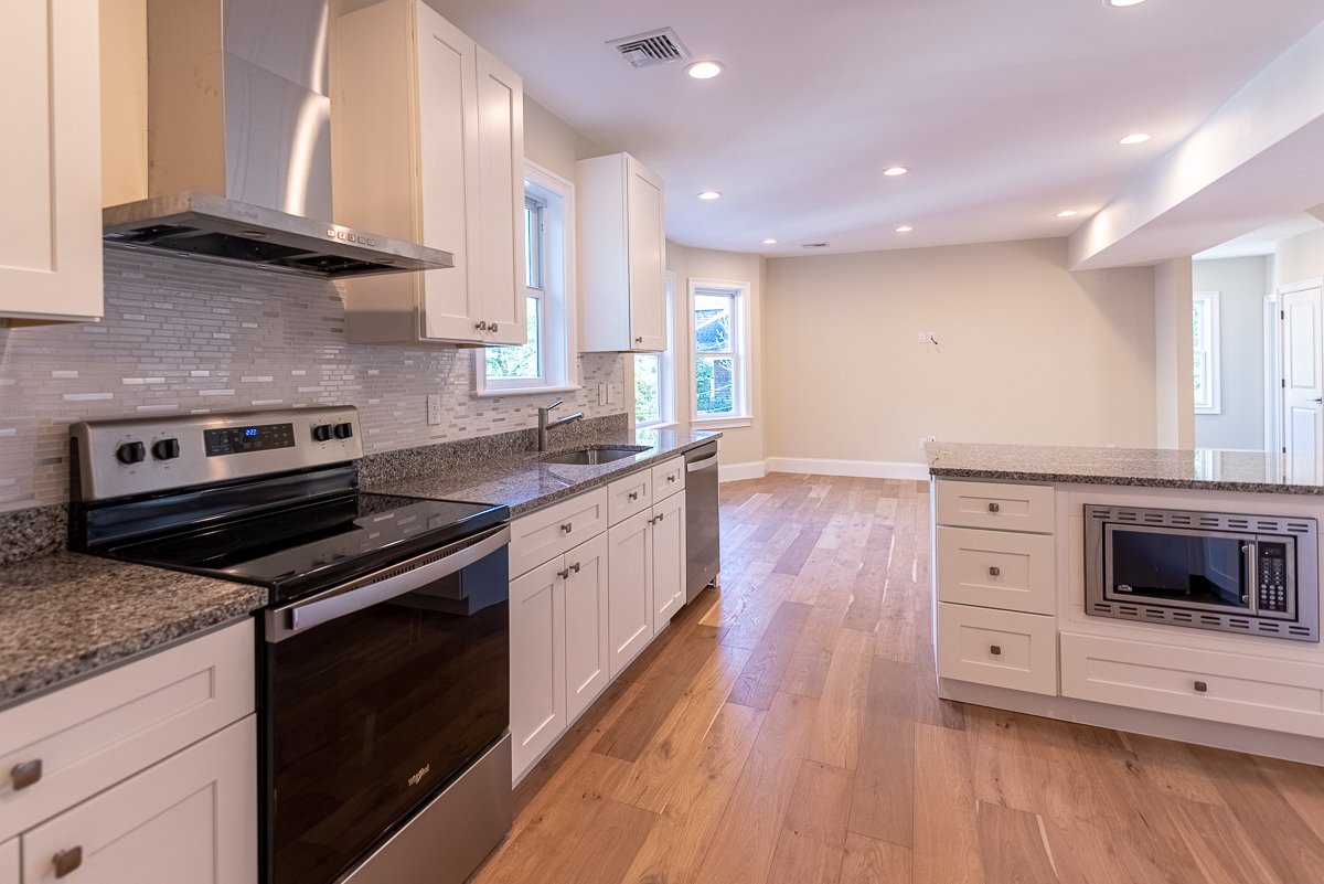 Empty modern kitchen with white cabinets, granite countertops, stainless steel stove and microwave, tile backsplash, hardwood floors, and large windows letting in natural light.