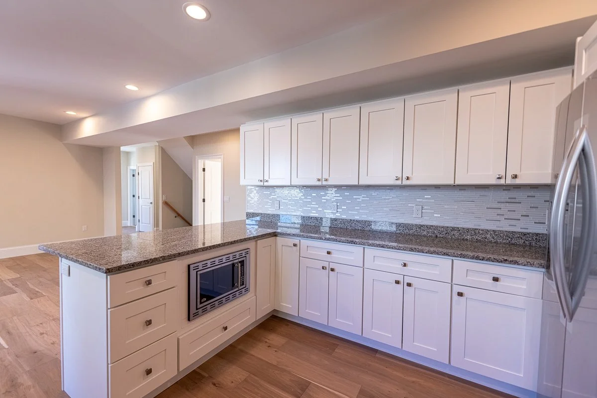 Modern kitchen with white cabinets, granite countertops, a stainless steel microwave, and a patterned tile backsplash. Hardwood flooring is visible.
