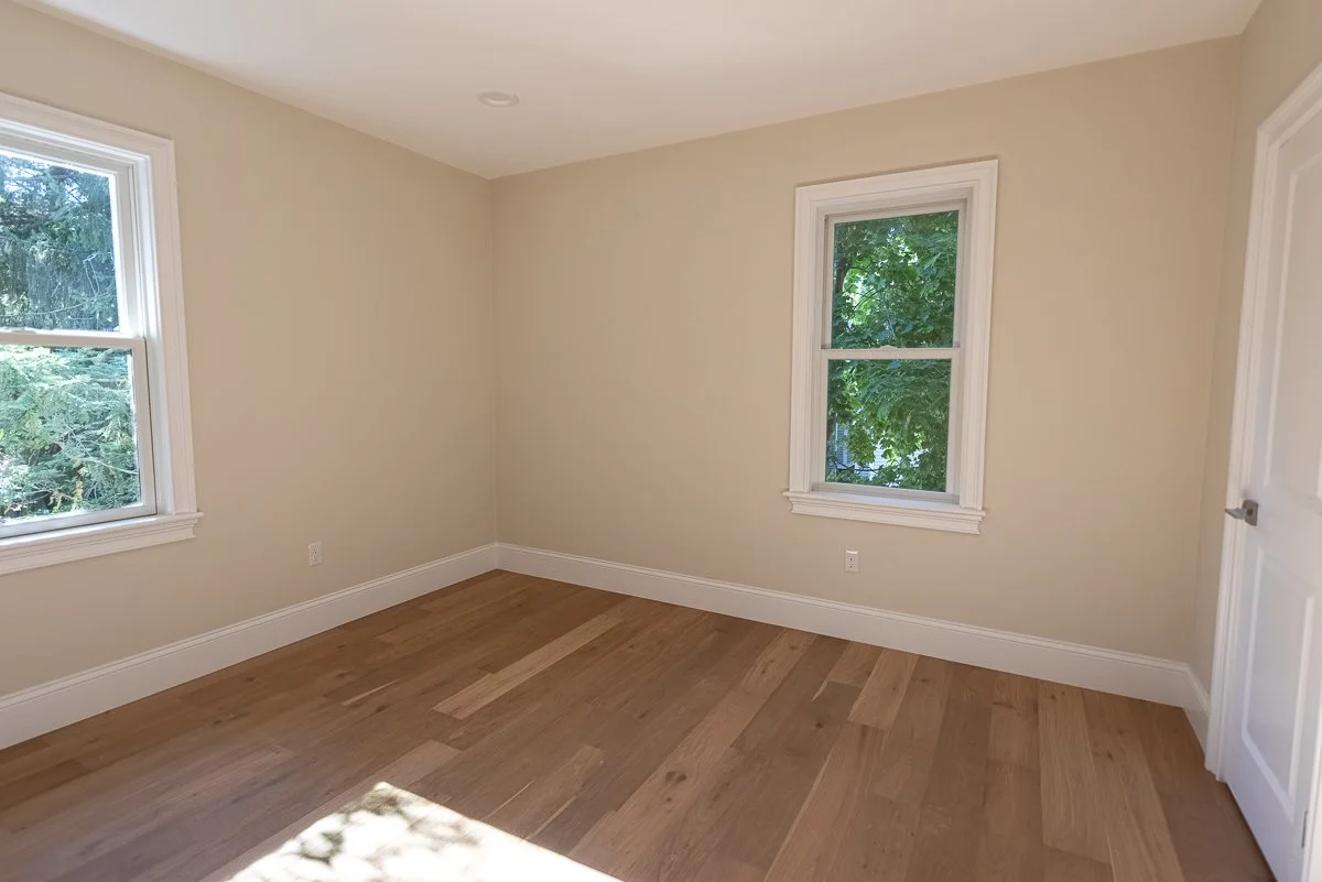Empty room with beige walls, hardwood floor, two windows with white trim, and a door.