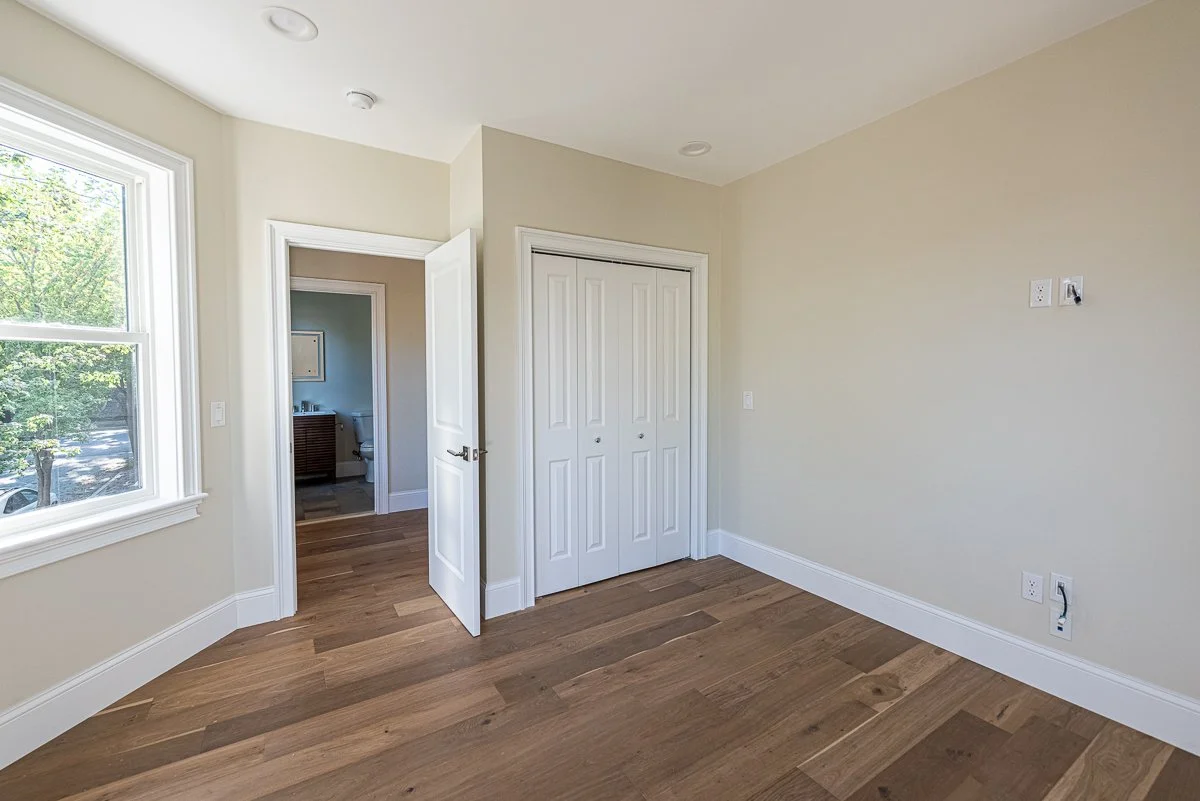 Empty room with hardwood floors, a large window, a small closet, and an open door leading to a bathroom.